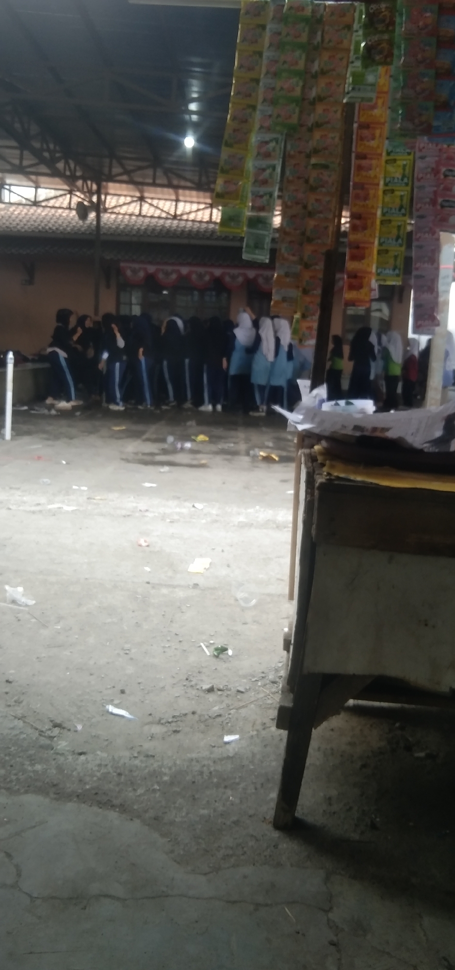 A group of approximately fifteen individuals, mostly young women dressed in dark headscarves and long-sleeved tops with blue stripes on their pants, are gathered in what appears to be an outdoor market or stall area. Some are hunched over, suggesting they are looking at items or interacting with each other. They are positioned under a covered structure with a corrugated metal roof supported by visible beams. In the foreground, a wooden stall with a weathered surface and a light-colored top is partially visible, positioned to the right of the frame. The ground is unpaved, appearing to be a mix of dirt and concrete, with scattered debris such as wrappers and plastic bottles. Hanging from the overhead structure are rows of colorful, small packaged goods, possibly snacks or instant food products, which are arranged in vertical columns. The overall impression is of a busy, informal commercial setting. The scene is located in the city of Rahayu, Indonesia.