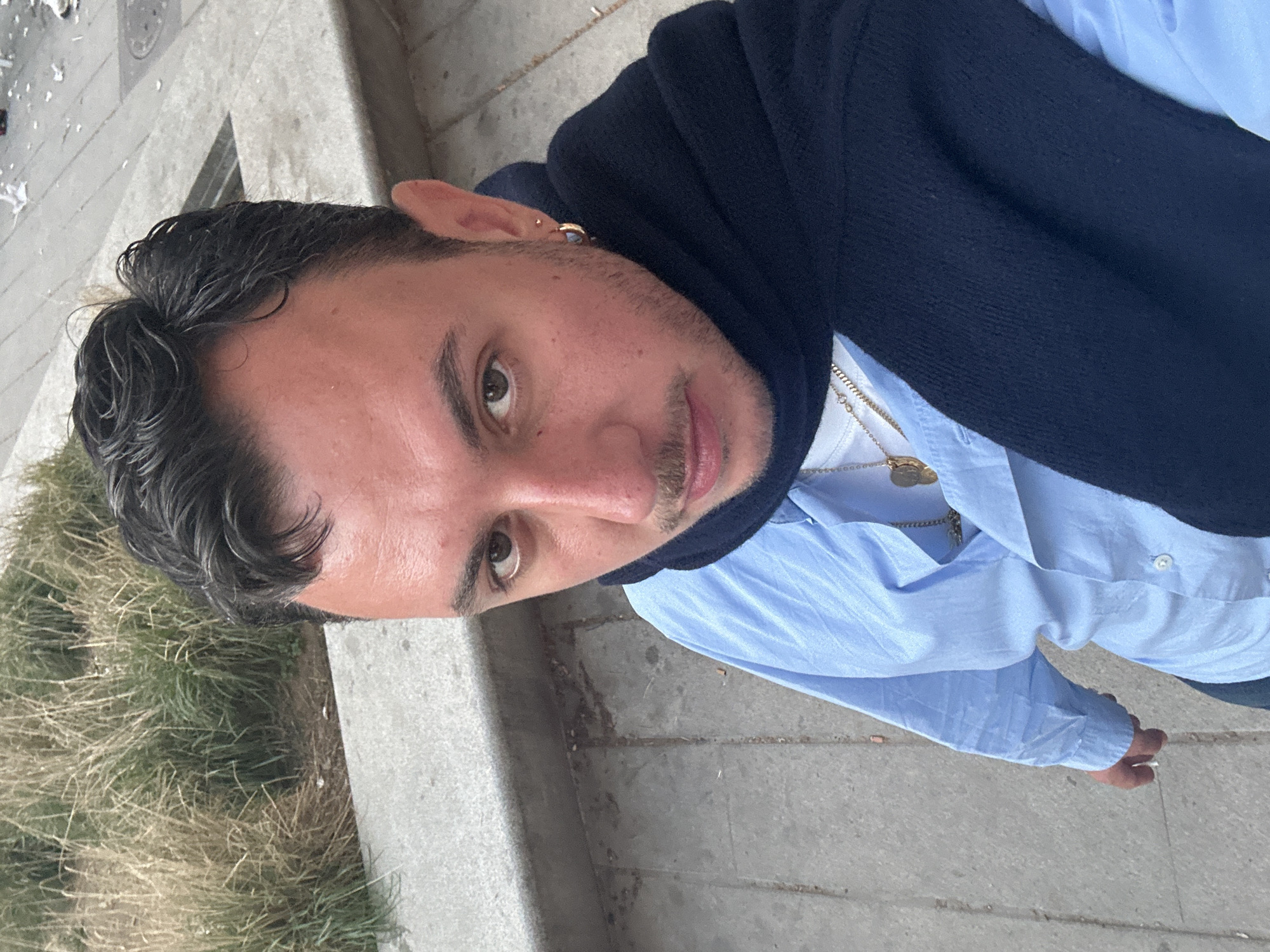 A man poses for the camera outdoors. He has short dark hair, light skin, and a mustache. He is wearing a light blue button-down shirt, a navy sweater draped over his shoulders, and a gold chain necklace. He has a gold earring in his left ear. The background includes a grassy area, a concrete walkway, and what appears to be the edge of a building. The location is suspected to be within the city of Madrid, Spain.