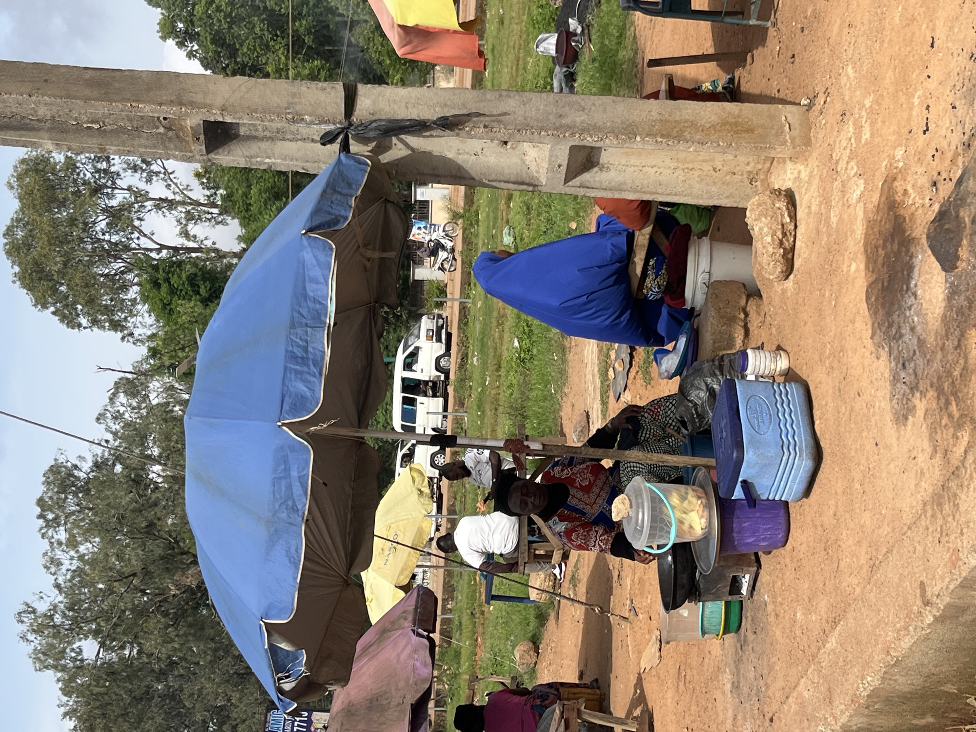 In the city of Bukuru, Nigeria, an outdoor market scene unfolds under a large blue umbrella. The ground is a dusty, reddish-brown earth, with patches of dry grass and scattered rocks. A concrete pillar, weathered and cracked, stands prominently to the right, secured by a black tie.
Several people are present, mostly seated. In the foreground, a woman wearing a red and blue patterned dress and a black headscarf sits on the ground, looking down at a collection of items. In front of her are cooking utensils, including a black pot on a small stove, and a clear plastic bucket filled with yellow items. A blue cooler box rests beside her.
Behind her, a woman draped in a vibrant blue hijab sits on a wooden bench, her back to the camera. To the left, under a yellow umbrella, two men are seated at a table. One wears a white shirt and the other a grey t-shirt with a logo. Further to the left, partially obscured by the blue umbrella, a woman in a pink top is seated on a stool.
In the background, a white minivan is parked. Trees with dense green foliage line the horizon, suggesting a warm climate. The sky is overcast, with a mix of blue and grey clouds. The overall impression is a bustling, everyday scene in a Nigerian marketplace.