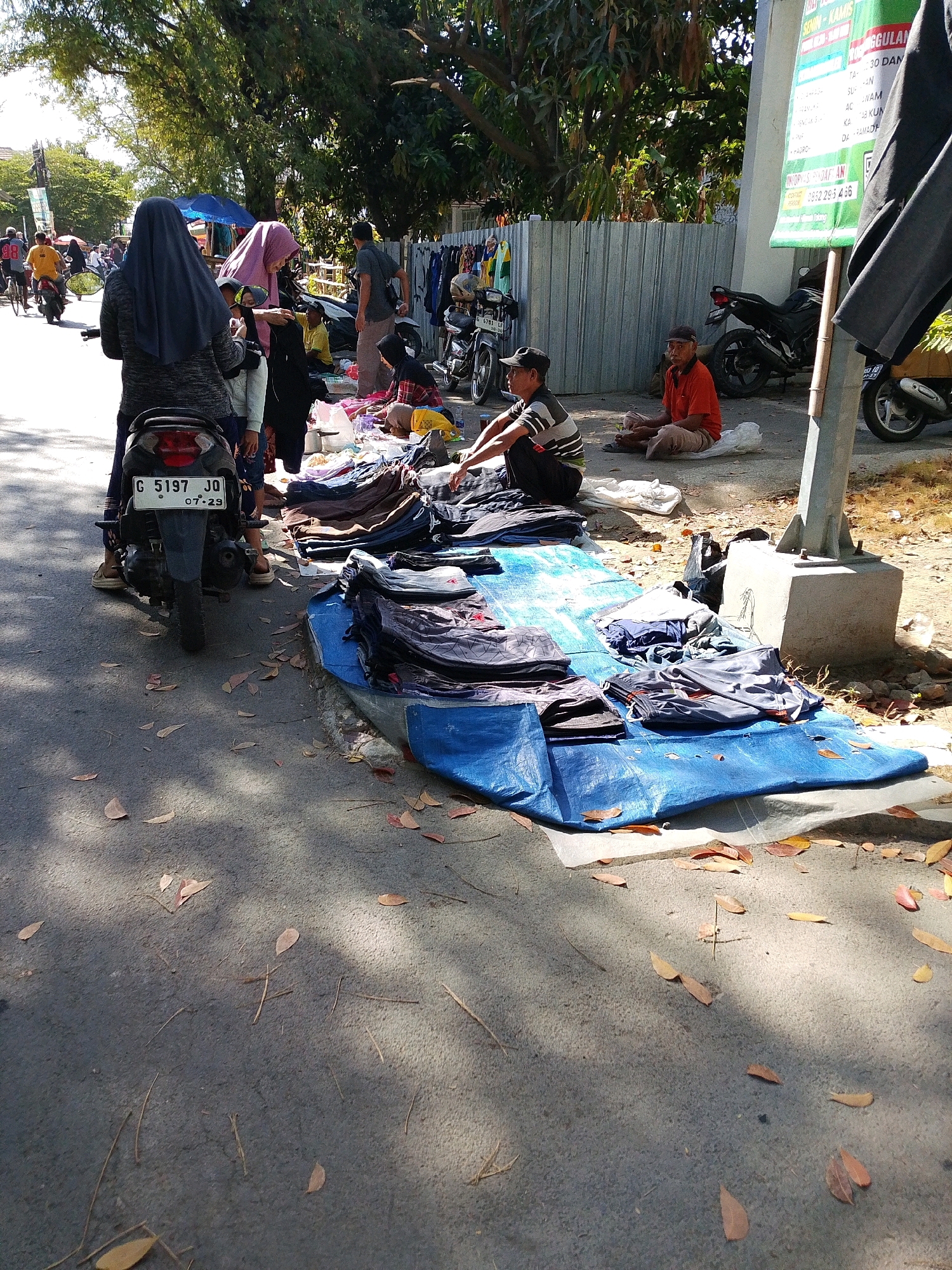 The image depicts a street-level scene in Tegalwangi, Indonesia, likely a market or roadside vendor area. In the foreground, a blue tarp is spread on the ground, displaying a collection of folded clothing, predominantly dark-colored pants and shorts. Several people are present, with varying degrees of engagement with the merchandise.
On the left side of the tarp, a person wearing a dark hijab and a patterned sweater stands near a motorcycle with license plate "G 5197 JQ". To their right, another person in a pink hijab is also present. A young child is visible between them.
Further back from the tarp, two men are seated on the ground. One man, wearing an orange shirt and light-colored pants, sits cross-legged. Another man, wearing a striped shirt and a dark cap, is also seated.
Behind the tarp and seated individuals, various objects are scattered, including more clothing items displayed on a fence-like structure and a motorcycle. A tall, gray pole with a concrete base is visible on the right side of the frame. A large, green banner with Indonesian text is attached to the pole. The environment appears sunny, with shadows cast by trees and possibly structures, and dried leaves are scattered on the ground. The background shows trees and a metallic fence.