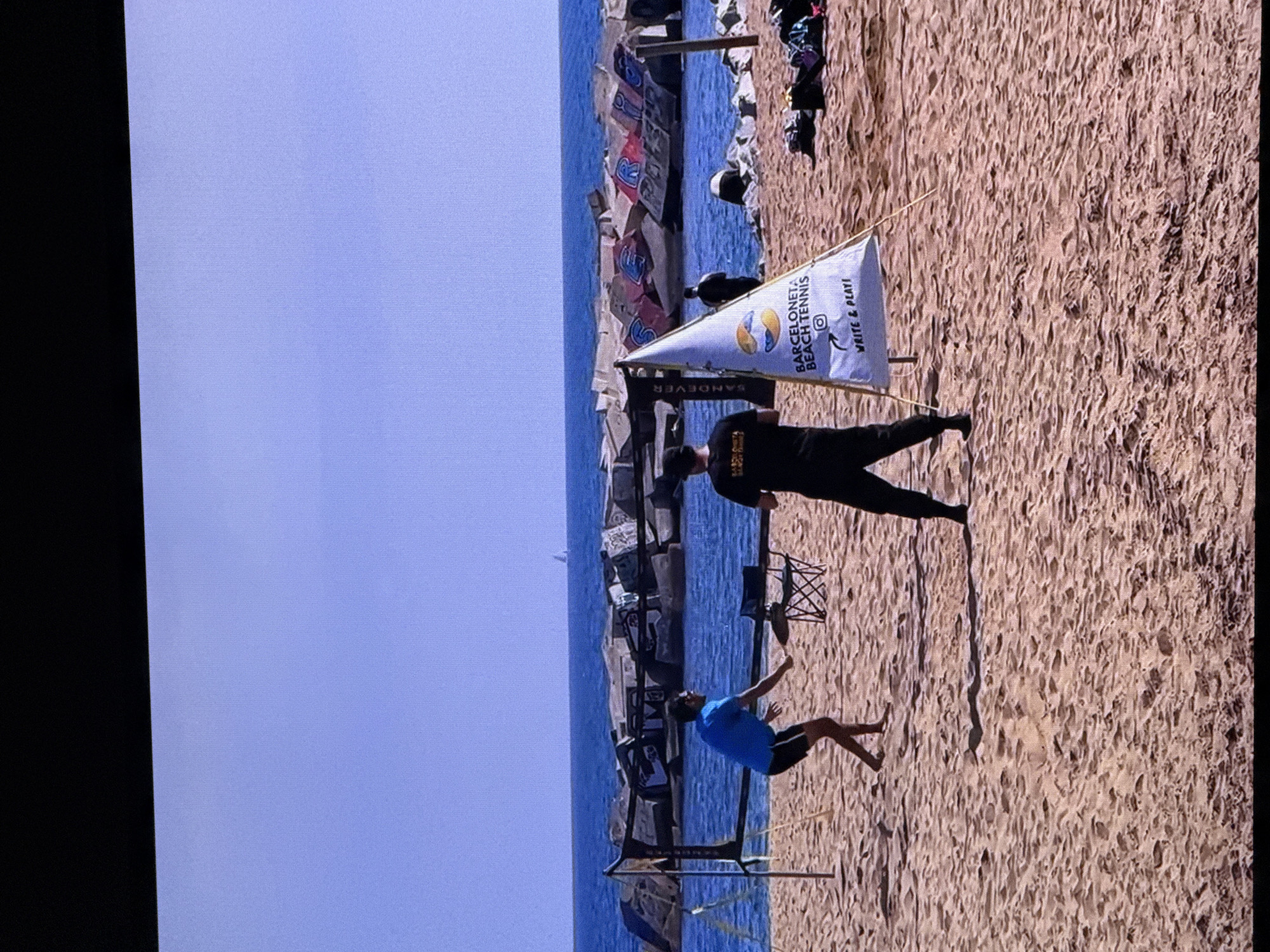This is a photograph taken on a sandy beach, likely in Barcelona, Spain, judging by the presence of a "Barceloneta Beach Tennis" banner. The scene depicts two individuals engaged in a beach tennis match. One person, wearing a blue t-shirt and black shorts, is mid-swing with a beach tennis racket. The other person, dressed in black, stands facing the net, also holding a racket. A net is set up between them, with poles on either side. In the foreground, a white triangular banner with the "Barceloneta Beach Tennis" logo and the tagline "WRITE & PLAY!" is visible. Behind the players, the Mediterranean Sea stretches out, bordered by a breakwater constructed from large concrete blocks, some of which are covered in graffiti. A small sailboat can be seen in the distance on the water. The overall atmosphere is one of recreation and sport in a coastal setting. The image does not depict Dubai, United Arab Emirates.