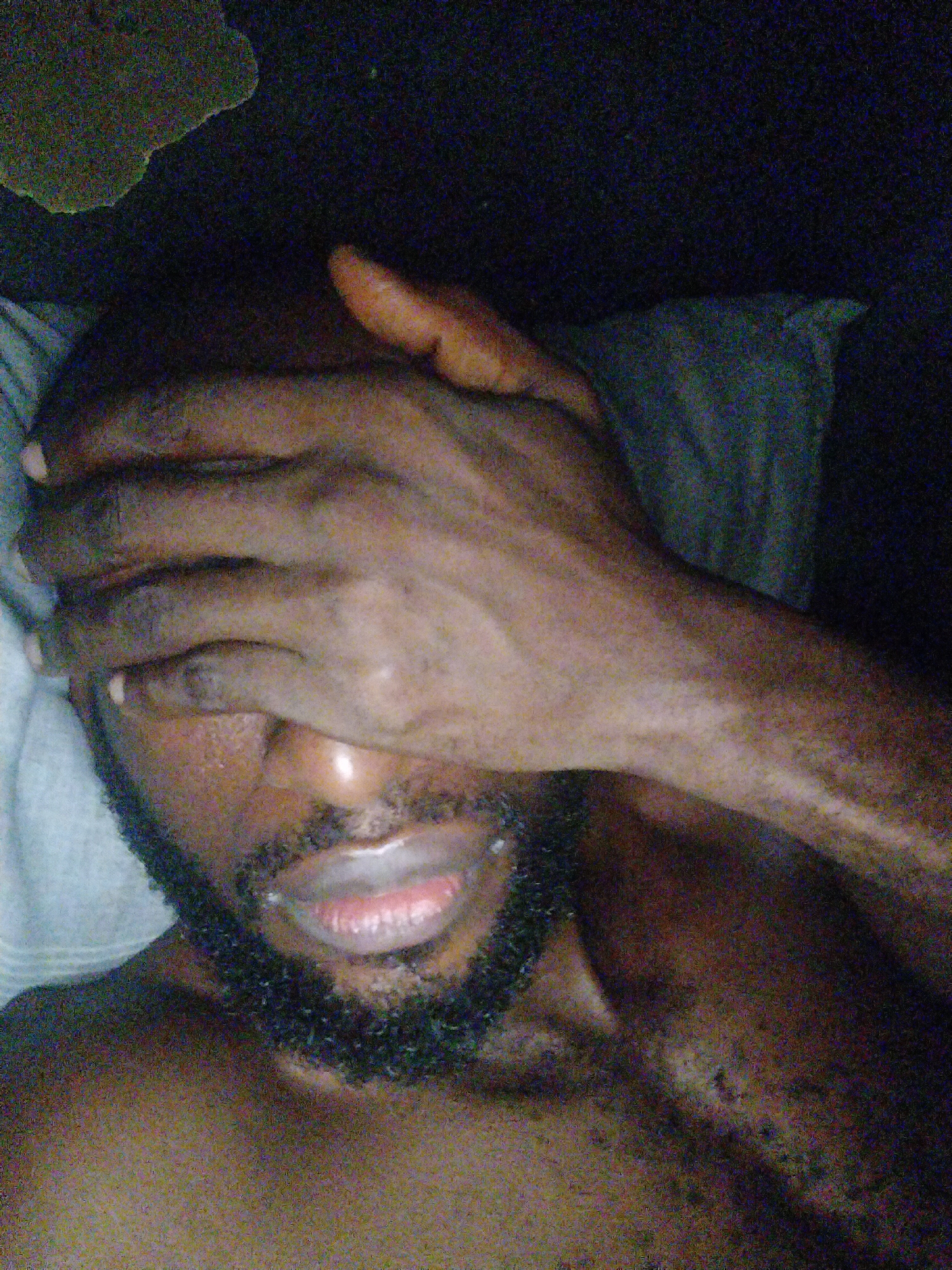 The image is a close-up selfie of a Black man lying down, covering his face with his right hand. His left arm is visible, resting on a pillow. The man has a beard and is wearing a light-colored shirt that is partially visible. The lighting is dim, with some areas of shadow and highlight. The background is dark, with hints of a pillow or bedding. The image appears to be taken indoors in a bedroom. There is no specific visual information in the image that would allow for the identification of the city of Uyo, Nigeria.
