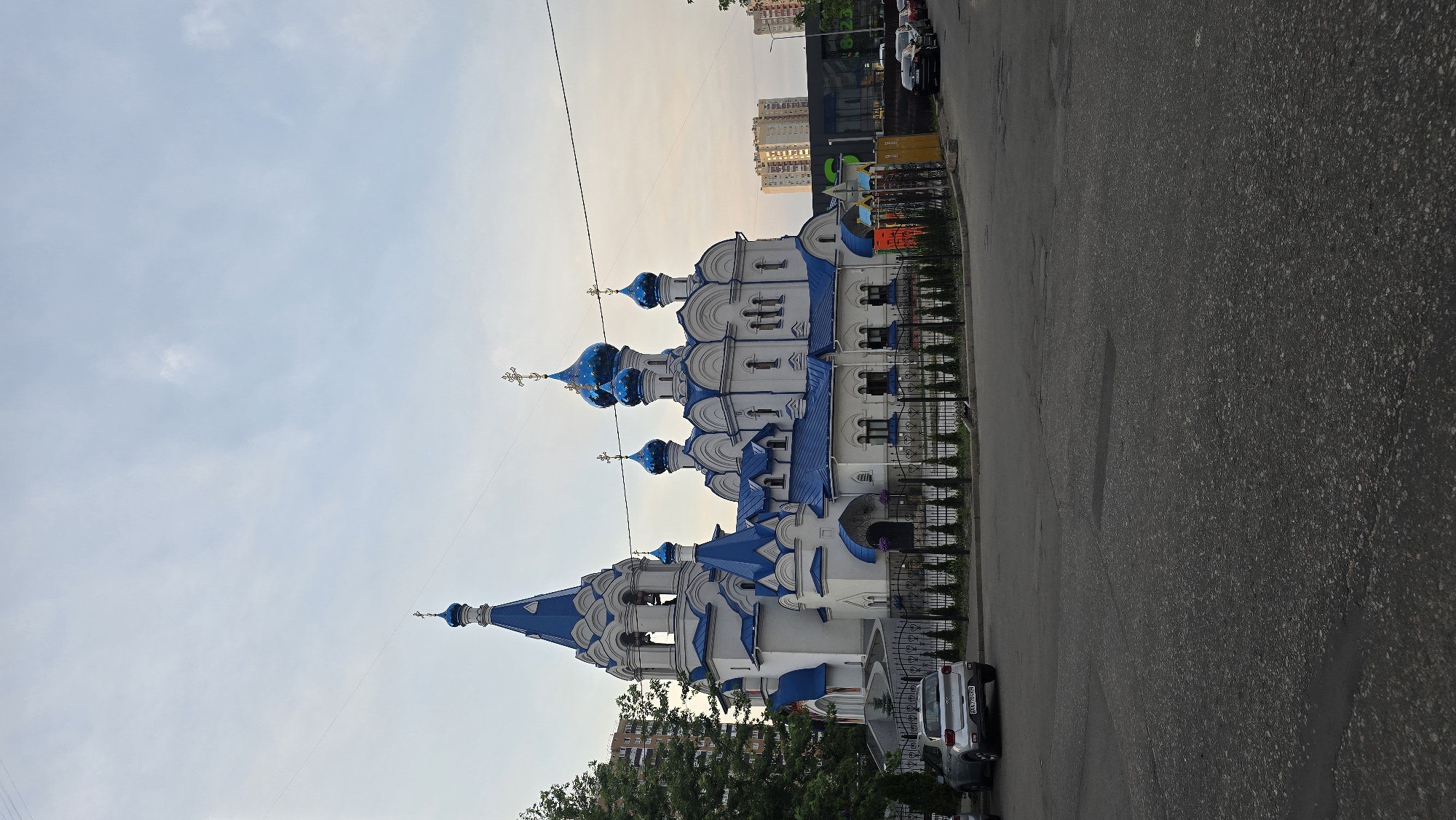 A white orthodox church with blue domes and a bell tower stands on the left side of the frame. A black metal fence encloses the church grounds. The church is situated on a paved street with a few parked cars. In the background, tall apartment buildings are visible. The sky is cloudy with a mix of grey and pale yellow hues.