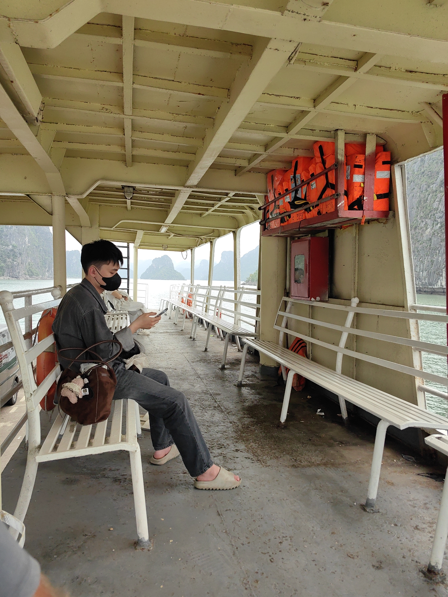 A person wearing a black face mask, grey denim shirt, and grey jeans sits on a white bench on a ferry. They are holding a phone and a brown handbag is next to them. Life jackets are stored above a red cabinet. The background shows limestone karsts in the water. The ferry is likely located in Bai Chay Ward, Vietnam.