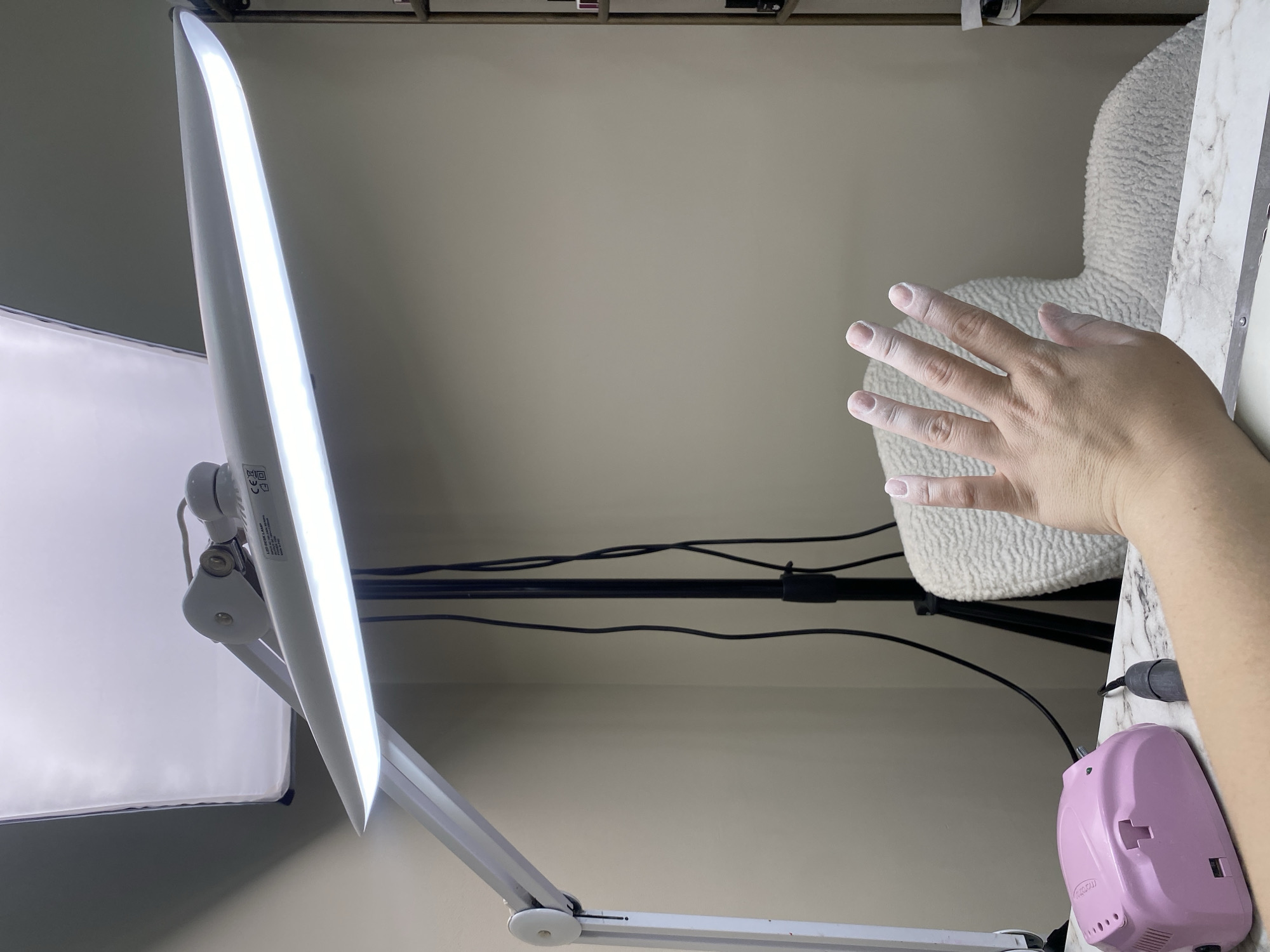 The image shows a close-up, slightly high-angle shot of a manicure setup. A white, articulated lamp with a large, flat, bright LED panel is positioned to the left. To the right, a person's hand is extended towards the center, with white powder or residue on the fingers. The background includes a cream-colored wall, some black cables, a fluffy white chair, and a marble-patterned surface. A pink electrical device is in the lower right corner, possibly a nail dryer or drill.
There is no information to suggest this image was taken in Sochi, Russia.
