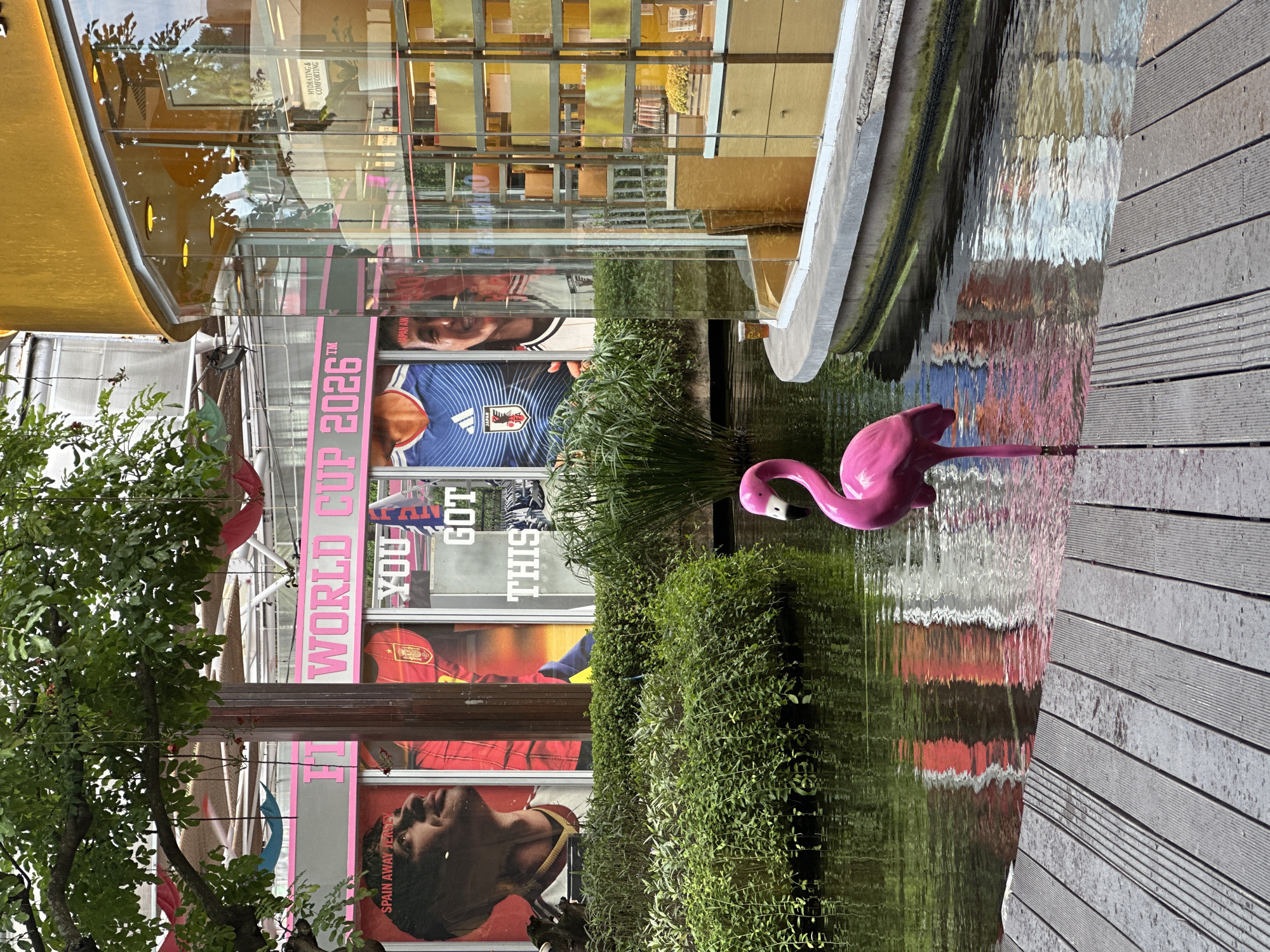 The image depicts a scene in Kuta, Indonesia, featuring a small artificial pond with a pink flamingo decoration standing in the water. The pond is adjacent to a wooden walkway. Behind the pond, there is a building adorned with large promotional banners for the FIFA World Cup 2026, featuring images of soccer players in their national team jerseys, specifically Japan and Spain. The building's architecture is modern with large glass panels, reflecting the surrounding environment and creating a distorted, mirror-like effect. The scene is partially obscured by greenery, including bushes and a tree, which contribute to the naturalistic feel of the otherwise urban setting. The overall impression is that of a commercial area with an attempt to incorporate natural elements for aesthetic appeal.