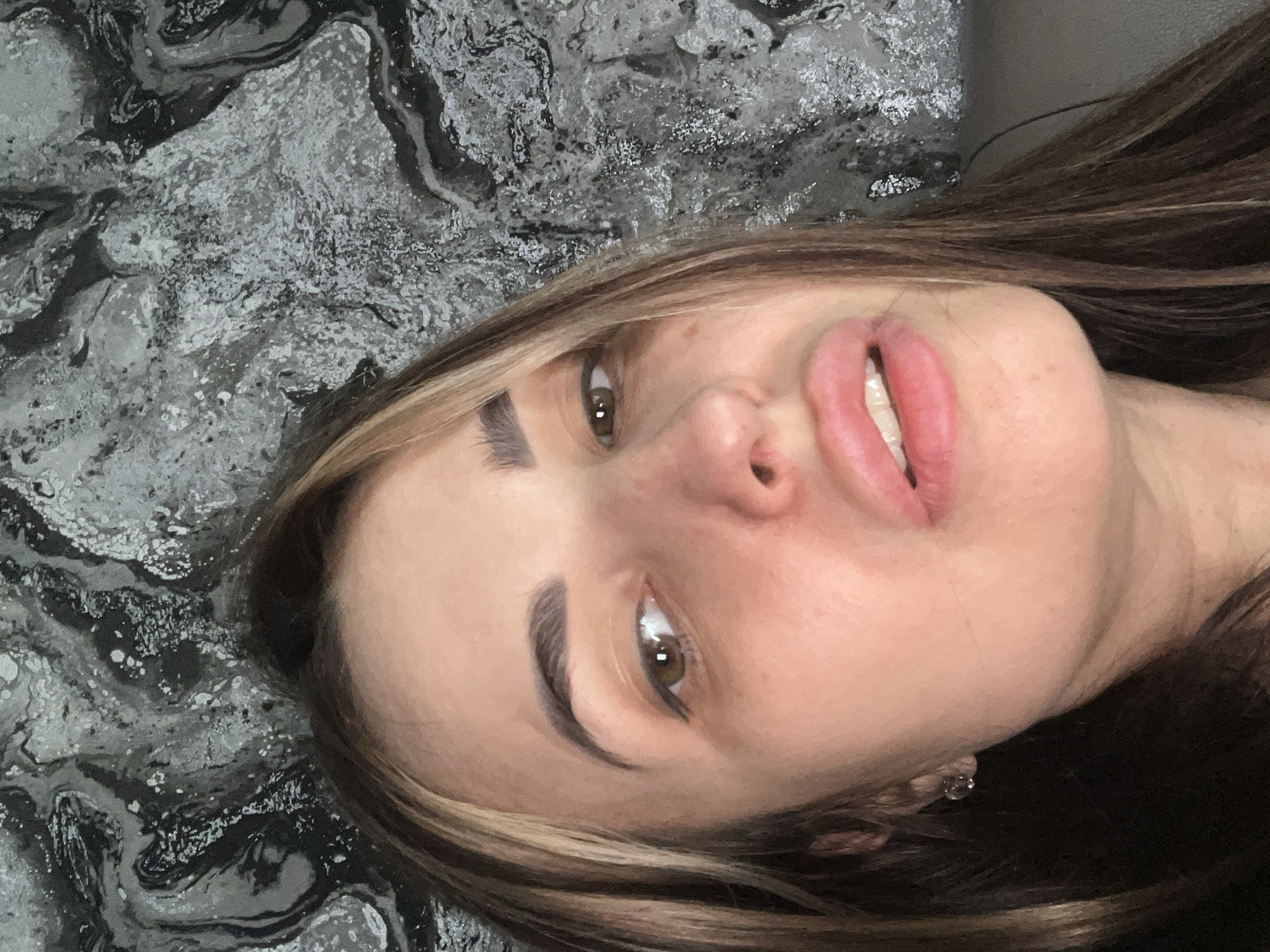 The image is a close-up selfie of a woman with light skin, brown hair with blonde highlights, and brown eyes. She is lying down on a surface with a gray and black marbled pattern. She is wearing an earring. The location is Unknown, Unknown.