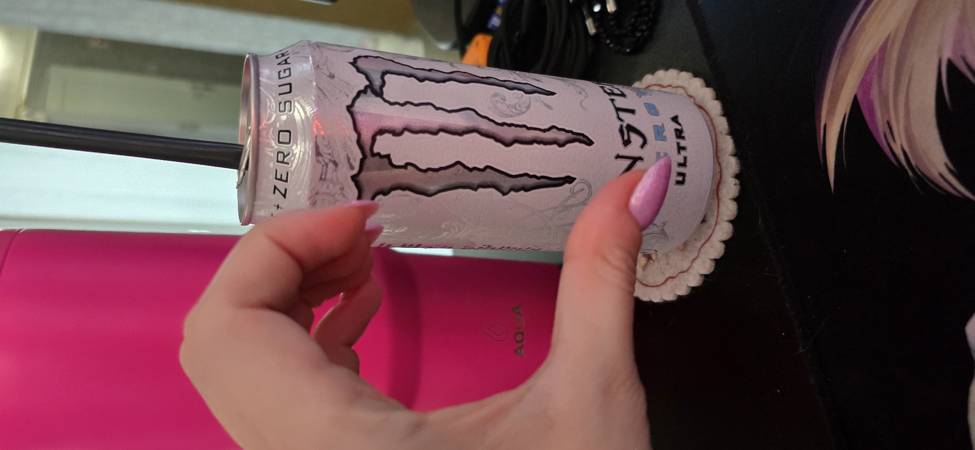 A hand with purple-painted fingernails is held in front of a white can of Monster Energy Ultra. The can is on a decorative coaster. Behind the can, a bright pink tumbler is partially visible. The scene appears to be indoors, possibly on a desk or table.
