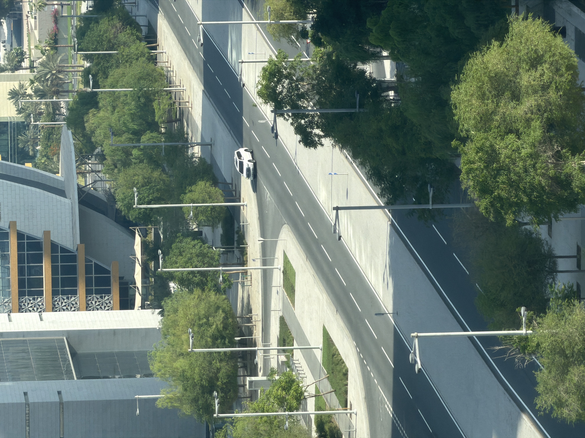 An aerial shot shows a modern city street scene with lush green trees bordering a road. A white SUV is driving on the road, and several streetlights are visible. The architecture of the buildings appears contemporary with large windows and geometric designs. The scene is bathed in daylight, casting shadows on the road and sidewalks.