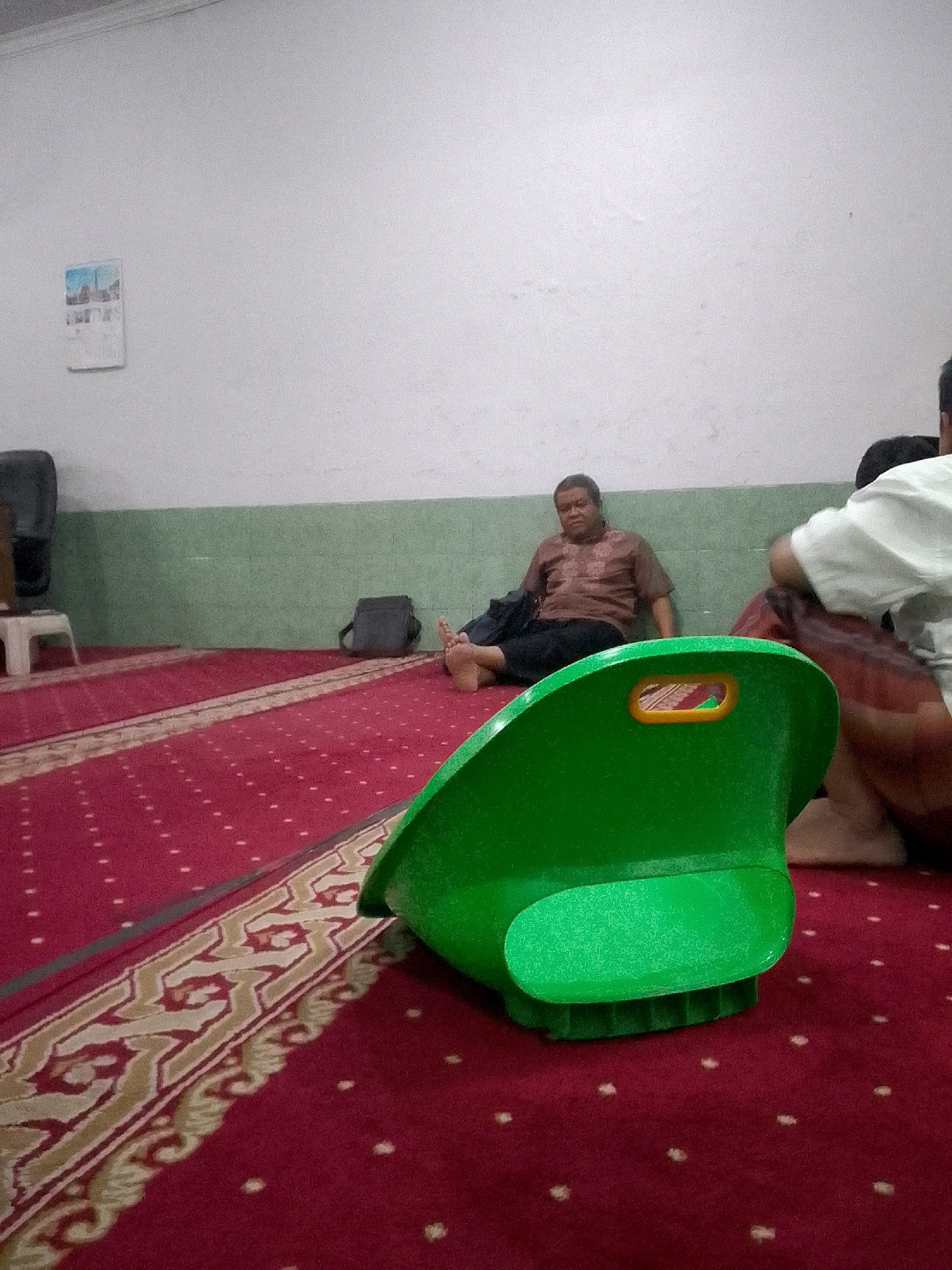 In Gunungmasigit, Indonesia, a man is seated on a red carpet in a room. He wears a brown patterned shirt and dark pants, with his legs extended and bare feet visible. He is leaning against a light green wall, with a dark bag next to him. In the foreground, a bright green plastic chair with an orange opening is positioned on the carpet. Another person, partially visible from the back and wearing a white shirt, is also seated on the carpet to the right of the green chair. A framed picture hangs on the white wall above the seated man. The carpet has a red base with a repeating pattern of gold and cream borders and small white dots.