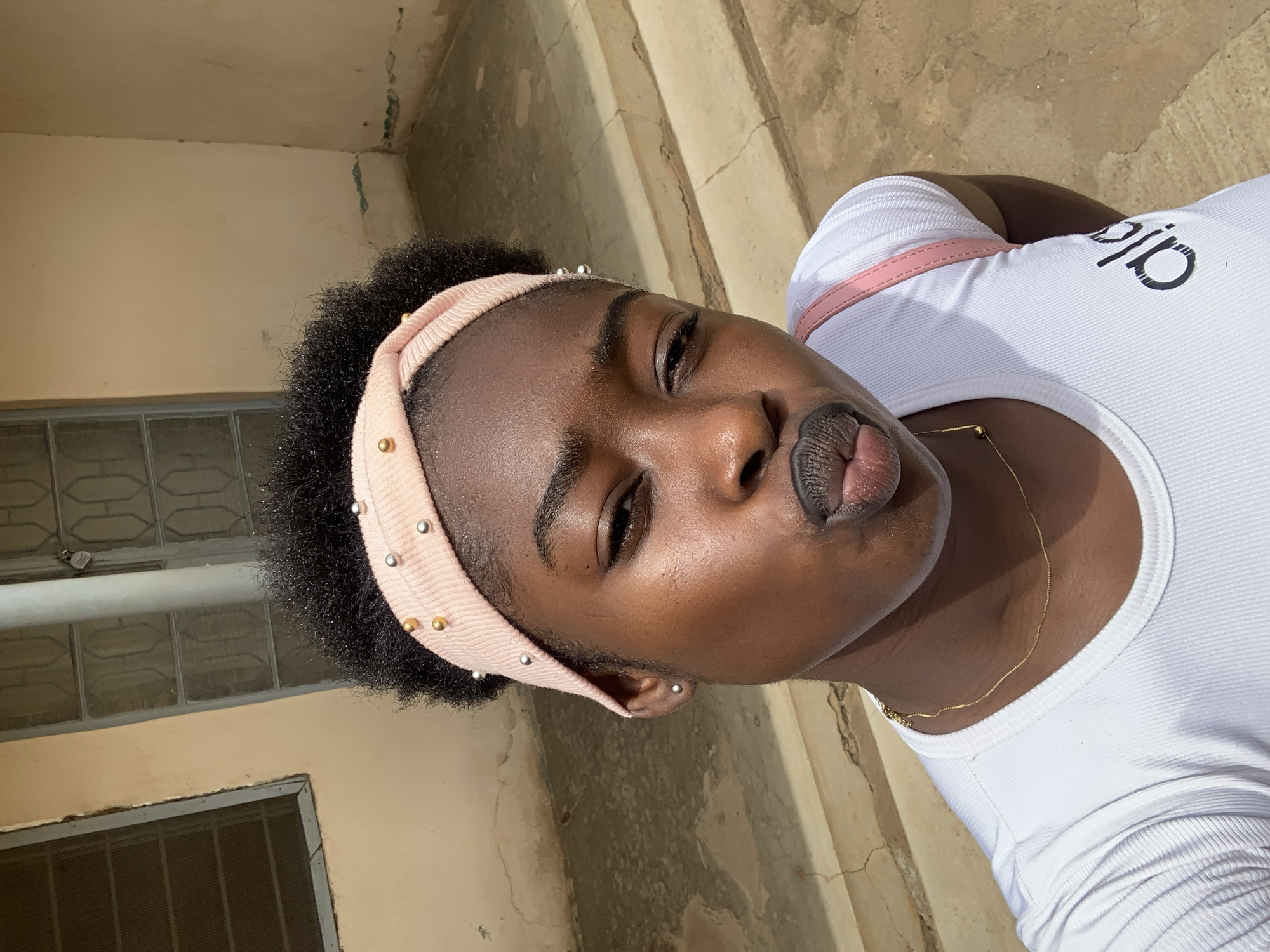 A young woman poses for a selfie outdoors. She has dark skin, short black hair under a light pink headband with silver and gold studs. She wears a white t-shirt and a delicate gold necklace. Her lips are pursed. The background features a beige wall with a window and textured concrete steps. The location is Rafin Par, Nigeria.
