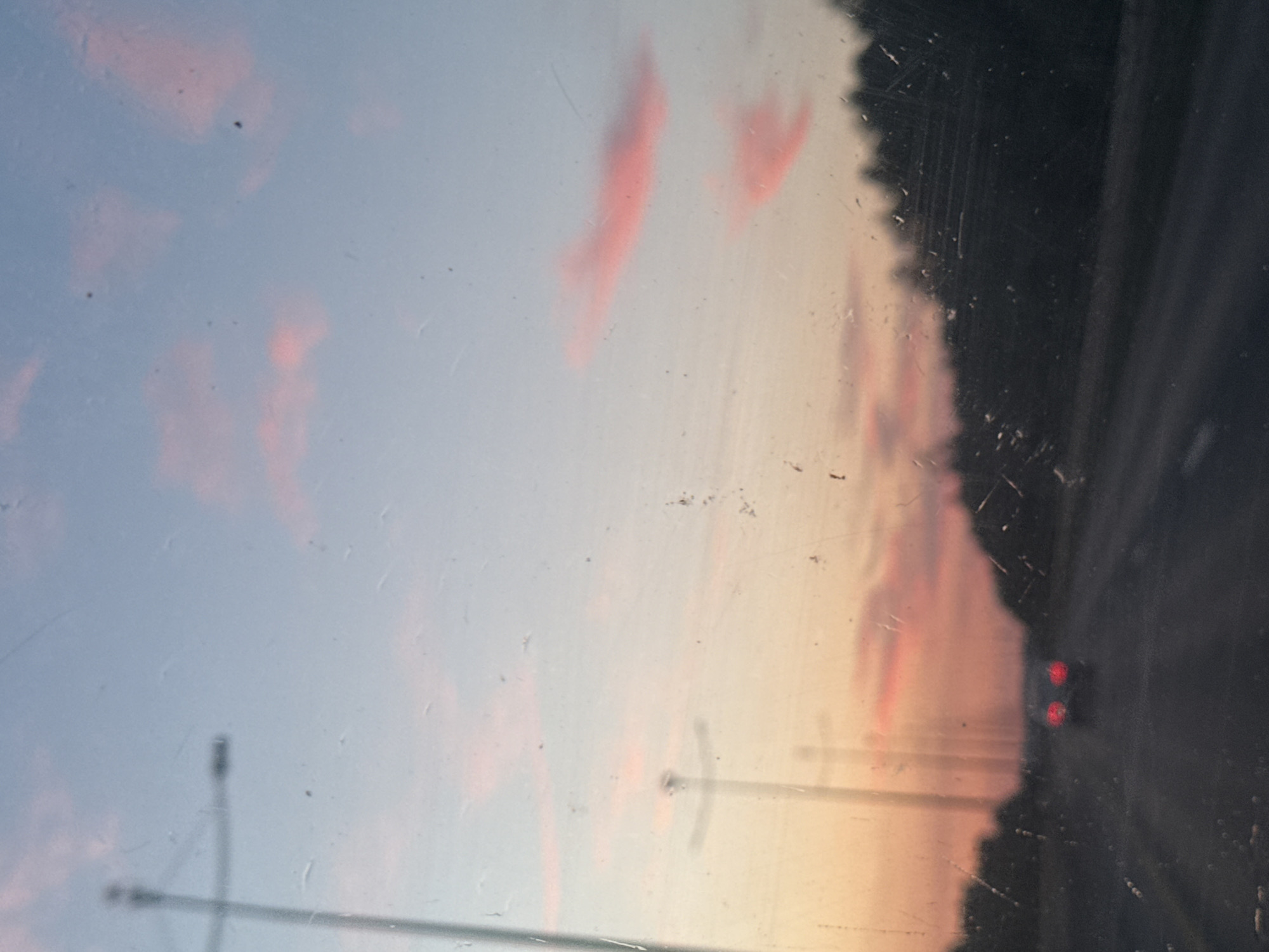 The image depicts a sunset over a road, likely taken from inside a vehicle. The sky is a gradient of light blue and pale orange, with wispy pink clouds scattered across it. The sun is setting, casting a warm glow on the horizon. A dark silhouette of trees lines the right side of the road, which appears to be an asphalt highway. In the distance, the red taillights of a car are visible, suggesting that traffic is moving along the road. The image is slightly blurred, and there are visible scratches and smudges on the surface from which the photo was taken, possibly a window. The overall mood is serene and peaceful, evoking the quiet beauty of a fading day. The location context provided places the scene in Kondratovo, Russia.