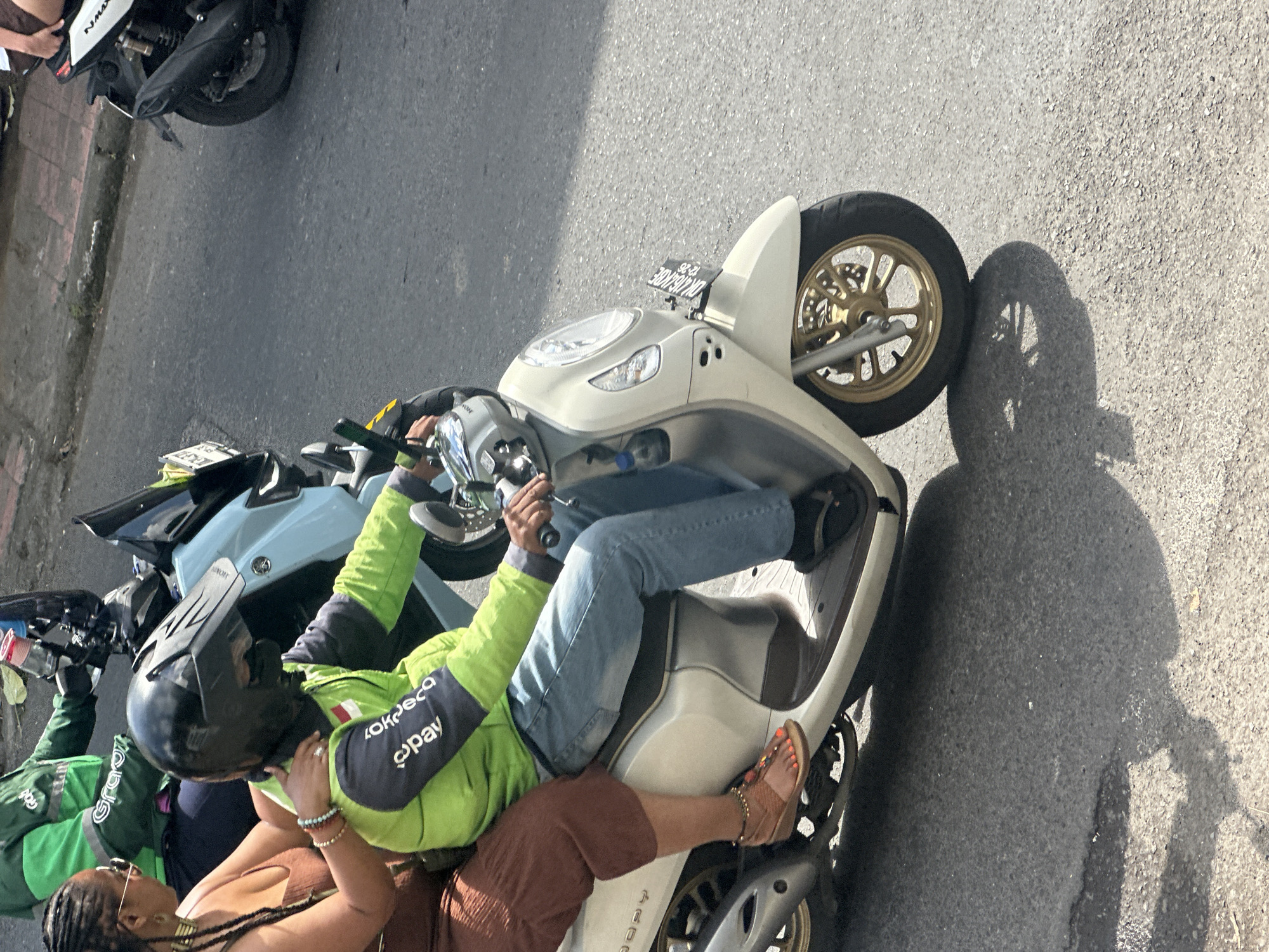 Two people are riding on a white scooter on a paved street in Ubud, Indonesia. The person in the front, wearing a black helmet and a green and dark blue jacket, is driving. The person behind them, wearing a brown dress and sunglasses, is a passenger. The scooter has a license plate that appears to read "DK 4763 RBE". The street is sunlit, and there is a shadow cast on the ground from the scooter and its riders. In the background, another scooter with a person in a green jacket is visible.