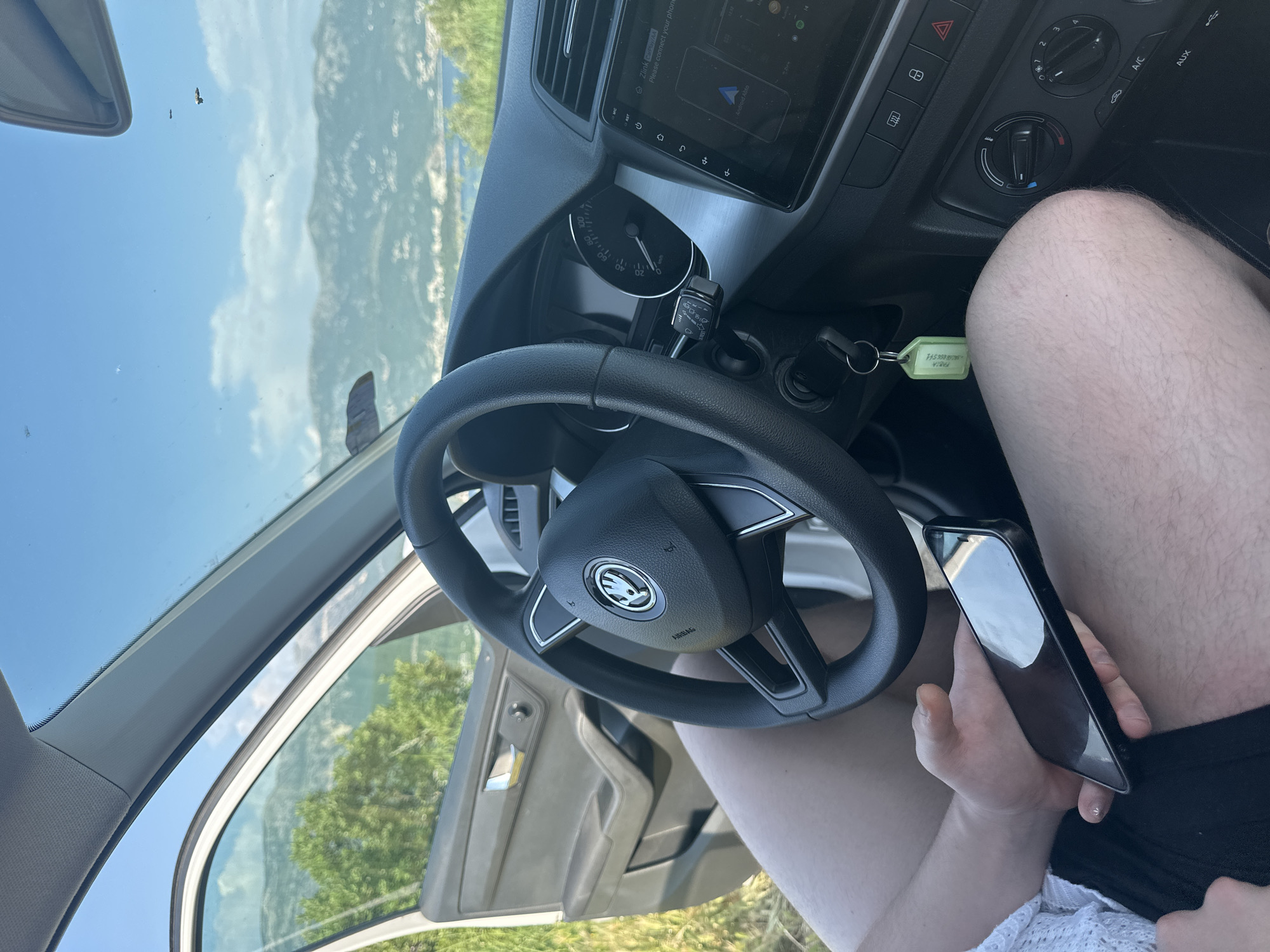 A first-person perspective image shows the interior of a Skoda automobile, with the steering wheel prominently displaying the Skoda logo. The car's dashboard features a media display screen and climate control dials. The driver, visible partially, holds a smartphone in their left hand. The driver is wearing black shorts and a white lace shirt.
Outside the open car door, greenery suggests a natural setting. Through the windshield, a mountainous landscape is visible, along with a blue sky. The text on the key chain indicates that the car may be in Budva, Montenegro.