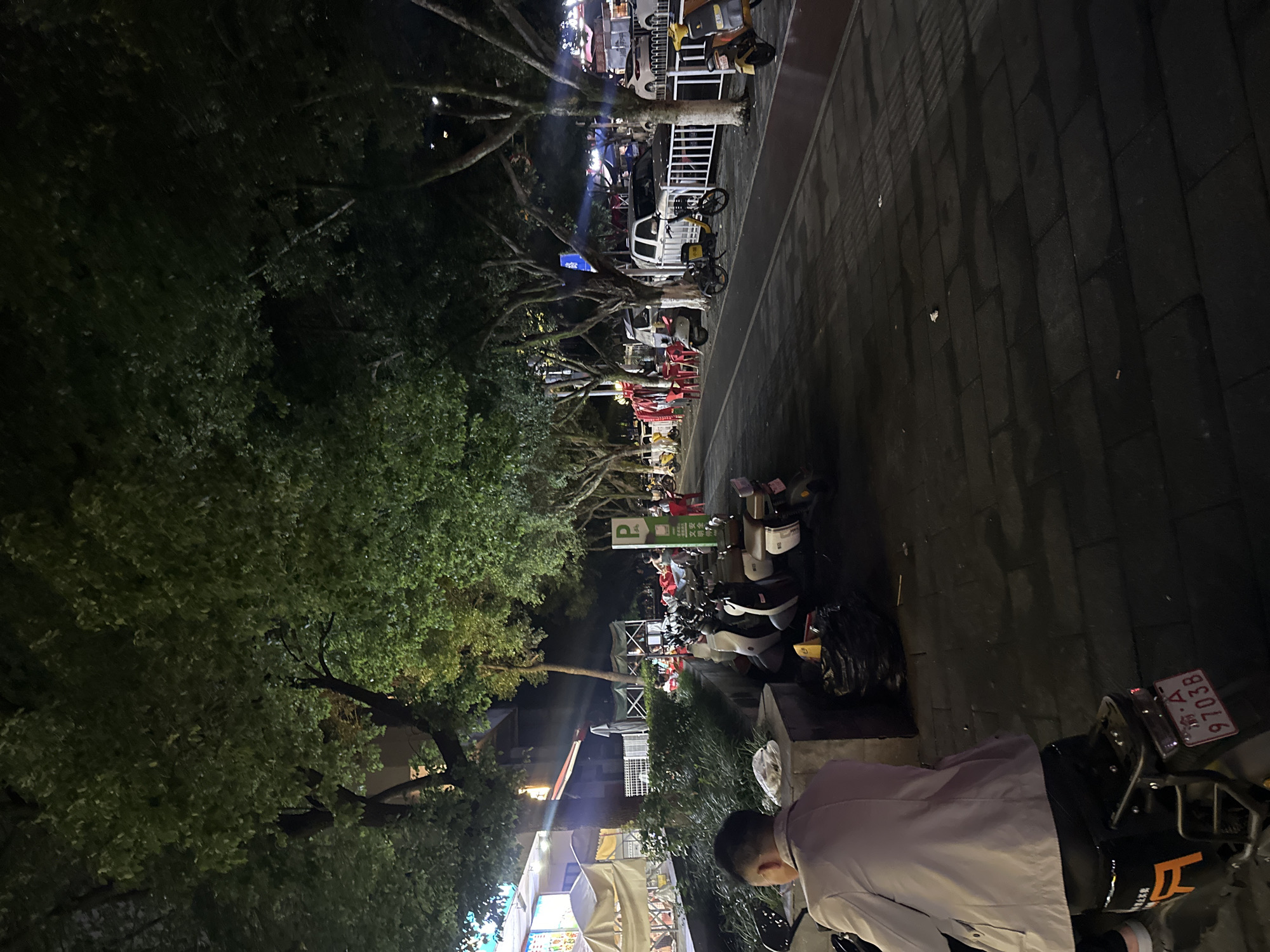 The image depicts a nighttime scene in Chongqing High-tech Zone, China. A tree-lined street is visible, with dense foliage on the left side. On the right, a paved walkway leads along the street. Parked motorcycles and scooters line the walkway. In the foreground, a person wearing a light-colored jacket is partially visible, standing next to a motorcycle. A license plate on the motorcycle reads "渝A 97038." Further down the street, various structures and signs are illuminated, including a "P" parking sign. Some businesses can be seen along the street. The lighting is dim, with light sources scattered throughout the scene.