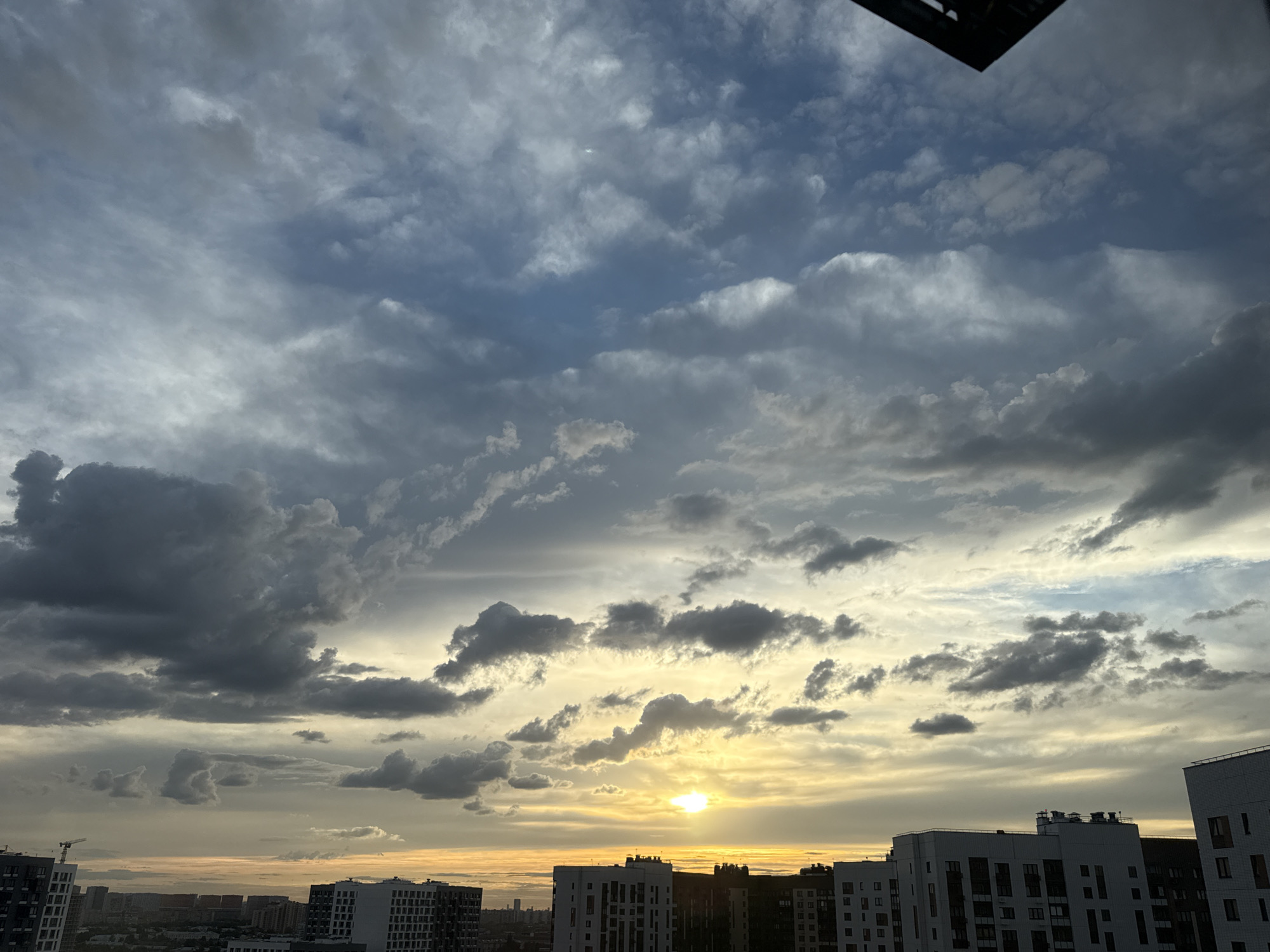The image depicts a cloudy sky over the city of Lyubertsy, Russia, likely during sunset or sunrise. The sky is filled with a mix of dark, heavy clouds and lighter, wispy formations, with a warm, golden light breaking through in the distance, suggesting the sun is low on the horizon. Below the sky, a cityscape is visible, characterized by numerous multi-story residential buildings, some appearing modern with white facades and dark windows. A construction crane is visible in the far left of the frame, indicating ongoing development in the area. The overall scene is urban, with a focus on the atmospheric conditions and the architectural landscape. There are no visible people or specific actions taking place.