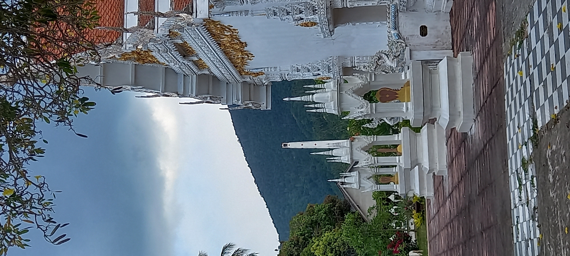 The image displays a serene temple complex nestled at the base of a lush, tree-covered mountain under a partly cloudy sky. In the foreground, a path features a distinctive pattern of brown tiles and black-and-white checkered sections, with scattered yellow petals or leaves. To the left, branches from a large tree extend into the frame, adding a natural element.
The mid-ground is dominated by ornate, white structures, indicative of traditional Thai Buddhist architecture. A large building with a reddish-orange tiled roof is visible, featuring intricate white gables and cornices accented with gold decorations. In front of this, several smaller, multi-tiered white chedis or stupas rise, characterized by their pointed finials and detailed carvings. A particularly tall, slender white stupa stands out prominently, reaching towards the background mountain. The intricate detailing on all the white structures highlights fine craftsmanship.
The background features a majestic, verdant mountain, its slopes covered in dense green foliage, rising steeply under a bright sky with patches of white clouds. The lighting suggests a daytime setting, possibly late morning or early afternoon, with soft shadows. No people or specific activities are depicted, giving the scene a tranquil and contemplative atmosphere. There is no visible text. The setting aligns with the provided context of Baan Chaloklum, Thailand, known for its natural beauty and religious sites.