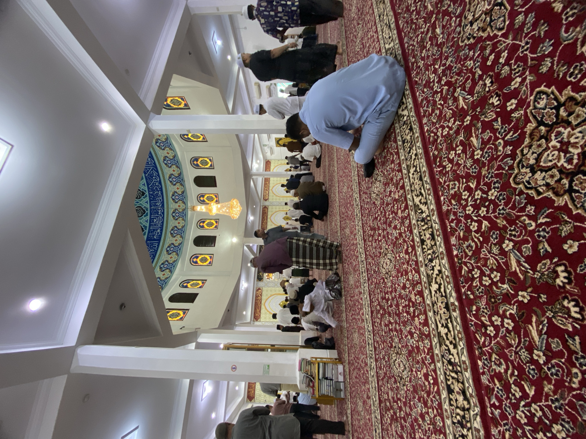 The image depicts the interior of a mosque, likely in Lhokseumawe, Indonesia, during congregational prayer. The foreground is dominated by a richly patterned red carpet with intricate floral designs. In the mid-ground, numerous men are seated or kneeling on the carpet, facing the front of the mosque. Many are wearing traditional Islamic attire, such as sarongs and caps. A prominent architectural feature is a domed ceiling adorned with Islamic calligraphy and colorful decorative elements. A chandelier hangs from the ceiling, illuminating the space. The walls are white, with arched windows featuring stained-glass designs. Pillars supporting the ceiling are also visible. The overall atmosphere suggests a peaceful and communal gathering for worship.
