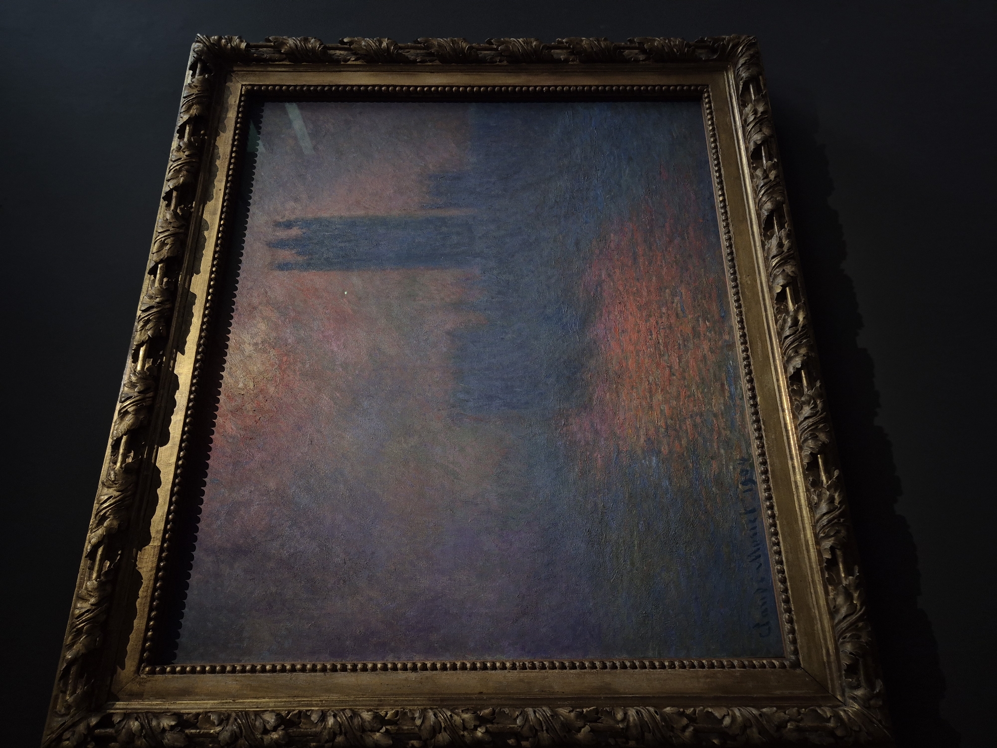 The image shows a framed painting of the Houses of Parliament in London, England, with the River Thames in the foreground. The painting is in an Impressionist style, with blurred brushstrokes and a focus on light and color. The sky is a hazy blend of pinks, purples, and blues, suggesting either sunrise or sunset. The architecture of the Houses of Parliament is rendered as dark silhouettes against the sky, with Big Ben appearing as a tall, slender tower. The river reflects the colors of the sky, creating a shimmering effect. The painting is signed "Claude Monet 1904" in the lower right corner. The frame is ornate and gilded, adding to the impression of a valuable artwork. The painting is displayed on a dark wall, which makes the colors of the artwork stand out.