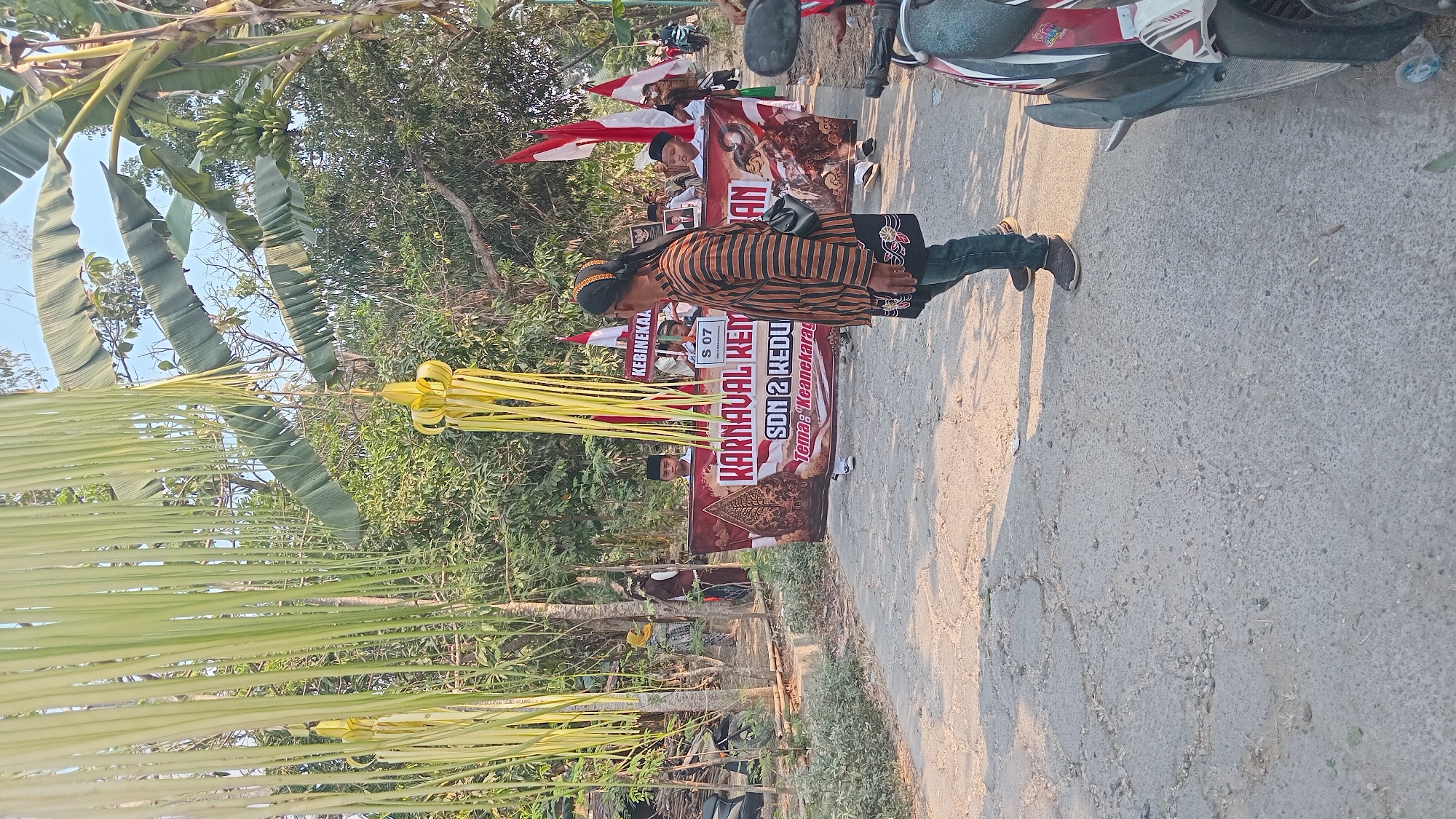 This image captures a street scene in Kedungwaru, Indonesia, during what appears to be a cultural parade or festival. In the foreground, a man dressed in traditional striped clothing and a headscarf stands facing away from the camera, looking towards a banner. Behind him, a large banner with the text "KARNAVAL KEM" and "SDN 2 KEDU" is displayed, suggesting it's an event organized by a local school. The banner also mentions "TEMA: Keanekaragam," meaning "Theme: Diversity." Several children in school uniforms are visible behind the banner, holding Indonesian flags.
The environment is rural, with trees and foliage lining the street, and banana leaves and a decorative yellow ornament hanging from above. The road is paved but appears worn and cracked. In the background, other people and a scooter are visible, indicating a public gathering. The lighting suggests it is daytime, with the sun casting shadows. The overall scene depicts a community event celebrating local culture and diversity in Kedungwaru.