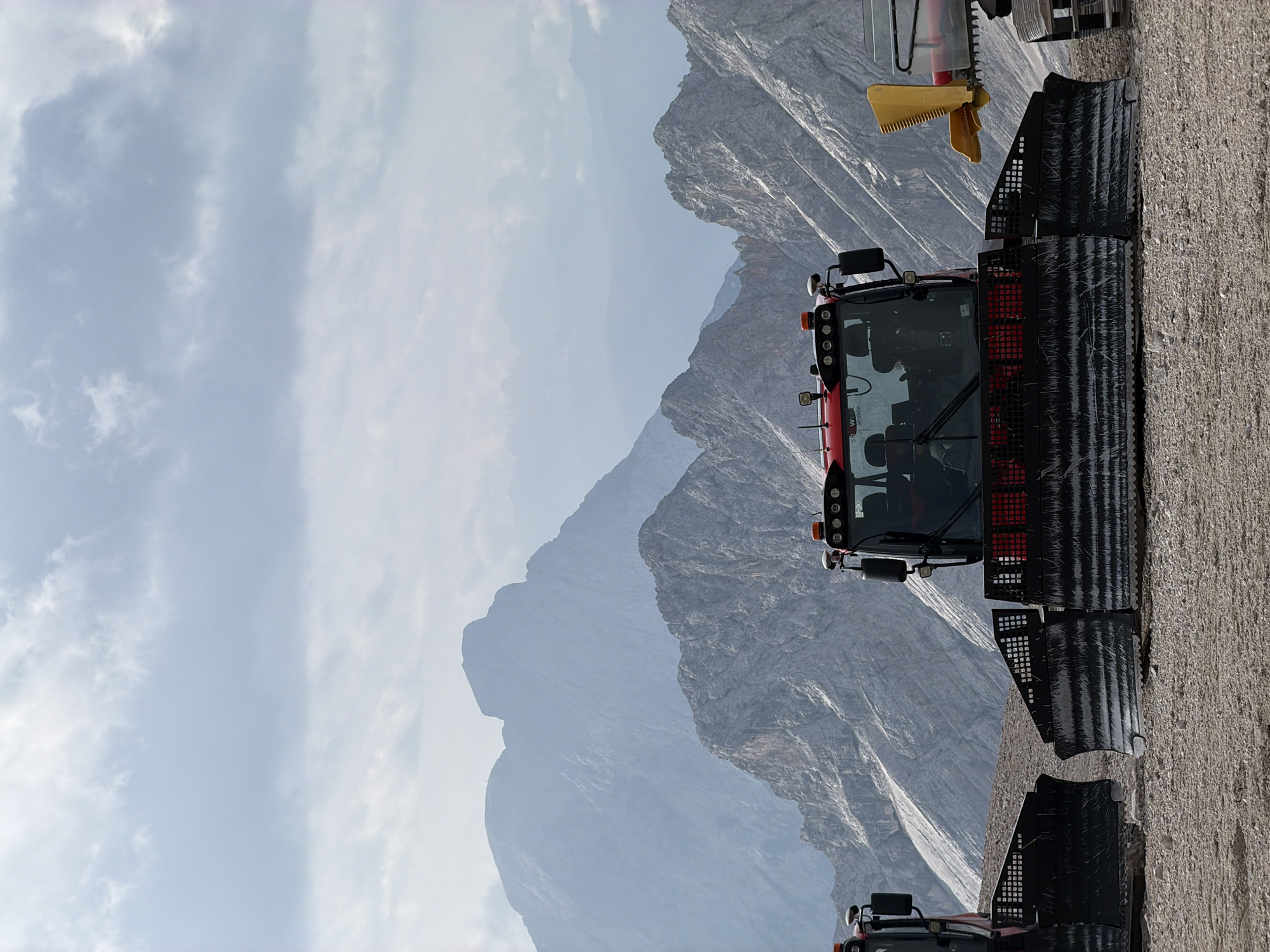 This image depicts a scenic mountain landscape in Garmisch-Partenkirchen, Germany, featuring two snow groomers parked on a gravelly terrain. The snow groomers, with their large plows, are positioned in the foreground, suggesting preparation for winter activities. Behind them, majestic, rugged mountains rise, partially obscured by atmospheric haze. The sky above is filled with layered clouds, creating a dramatic and expansive backdrop. The overall scene conveys a sense of natural beauty and the operational readiness of the mountain resort.