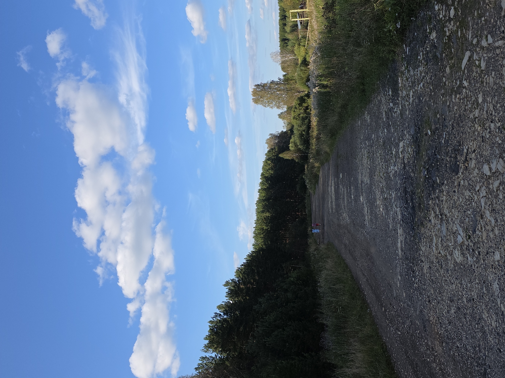 This image depicts a rural road in Magnitka, Russia, lined by a dense forest on the left and overgrown vegetation on the right. The road itself is unpaved and appears to be made of gravel and dirt, with some areas showing signs of wear and tear. In the distance, two figures, possibly children, are walking along the road, one in a pink garment and the other in a blue one. The sky above is a clear blue, dotted with fluffy white clouds, suggesting fair weather. The overall scene conveys a sense of natural serenity and peacefulness.