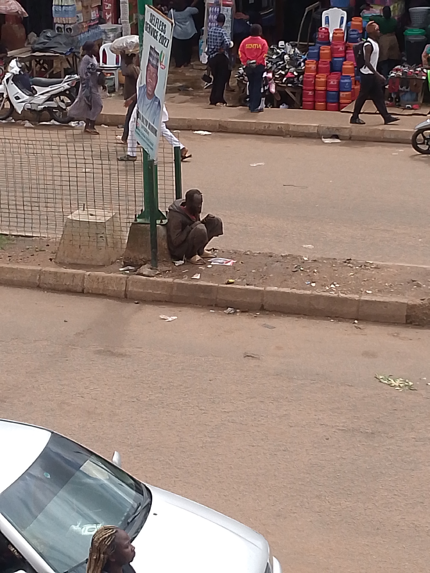 The image depicts a street scene in Bukuru, Nigeria. A person is sitting on the curb, hunched over and appearing to be looking at something in their hands. They are wearing dark clothing. The street is paved and appears to be a commercial area, with market stalls visible in the background. Several people are walking in the background, some carrying goods. A motorcycle is parked to the left, and a white car is partially visible in the foreground. A large sign is also present, featuring a picture of a man and text, likely a political poster. The environment appears somewhat cluttered, with litter visible on the ground.