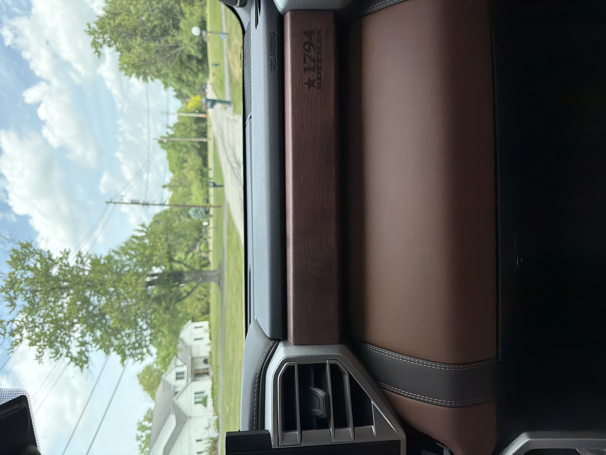 The image, taken from inside a vehicle, shows a view through the windshield of a suburban street scene in Pepper Pike, Ohio, United States of America. The foreground features the interior of the vehicle, with a brown leather dashboard and a silver air vent visible. A textured wood-grain trim with "1794 EDITION" and a star emblem is present on the dashboard. The middle ground shows a paved road lined with green trees and lawns. White houses with pitched roofs are visible on either side of the road, and mailboxes stand at the edge of the properties. In the background, power lines and utility poles are present against a cloudy sky. The overall impression is of a quiet, residential street on a bright, overcast day.