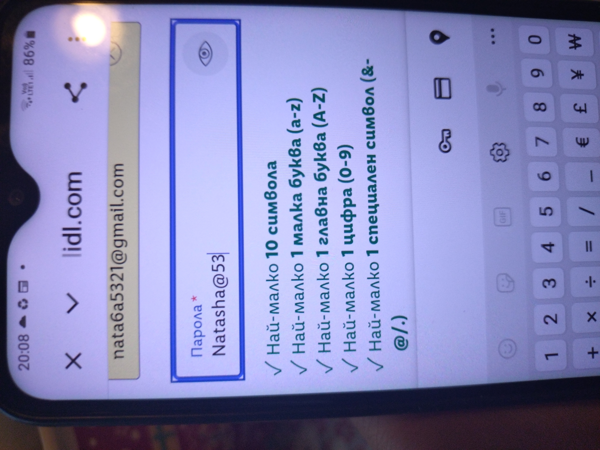 The scene depicts a close-up, slightly angled view of a smartphone screen, presumably in Stara Zagora, Bulgaria. The phone's display shows a web browser interface for "lidl.com." The status bar at the top indicates the time as 20:08, with mobile data (VoLTE LTE1) active, and battery life at 86%.
An email address, "nata6a5321@gmail.com," is displayed within an input field. Below this, a password input field, labeled "Парола *" (Password *), contains the partially entered text "Natasha@53." An eye icon on the right side of this field suggests a password visibility toggle.
Immediately beneath the password field, five password requirements are listed in Bulgarian, each preceded by a green checkmark indicating compliance. These requirements translate to:
* "Най-малко 10 символа" (Minimum 10 symbols)
* "Най-малко 1 малка буква (a-z)" (Minimum 1 lowercase letter (a-z))
* "Най-малко 1 главна буква (A-Z)" (Minimum 1 uppercase letter (A-Z))
* "Най-малко 1 цифра (0-9)" (Minimum 1 digit (0-9))
* "Най-малко 1 специален символ (&-@/.)" (Minimum 1 special symbol (&-@/.))
The password "Natasha@53" (10 characters) satisfies all listed criteria. The lower portion of the screen displays a virtual keyboard with numeric keys (1-0) and various symbols, including plus, multiply, divide, equals, forward slash, underscore, euro, pound, yen, and won. Key, folder, and location pin icons are also visible above the keyboard. A slight glare is present on the upper right side of the screen.