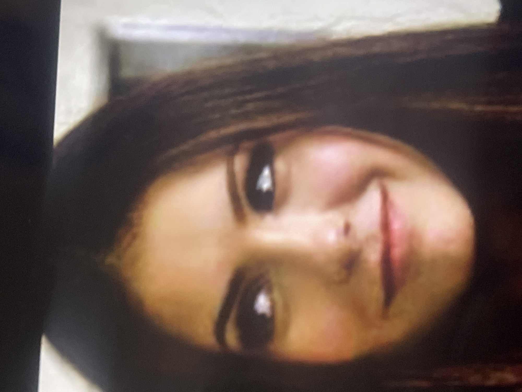 The image is a close-up, chest-up portrait of a young woman, rotated 90 degrees counter-clockwise. When viewed correctly, she is positioned centrally, looking directly at the camera with a subtle, soft smile or smirk. Her expression conveys a sense of calm or slight amusement.
She has long, dark hair that frames her face and dark, expressive eyes. Her complexion is fair, with noticeable rosy tones on her cheeks and around her nose, suggesting natural blush or specific lighting. Her eyebrows are well-defined and dark, complementing her hair color. A clear, bright reflection is visible in the pupils of both her eyes, indicating a light source directed towards her. Her lips are full, and her slight smile reveals the gentle curve of her mouth.
The setting appears to be indoors, likely a room. The background is largely out of focus, consisting of blurry light tones on the right side of the frame (behind her left shoulder) and a darker, indistinct vertical shape on the left (behind her right shoulder), possibly part of furniture or a doorway. The lighting is consistent with an indoor environment, illuminating her face clearly while casting the background into soft blur. There are no specific cues within the image to identify the location as Poltava, Ukraine, beyond the given context. No text is visible in the image.