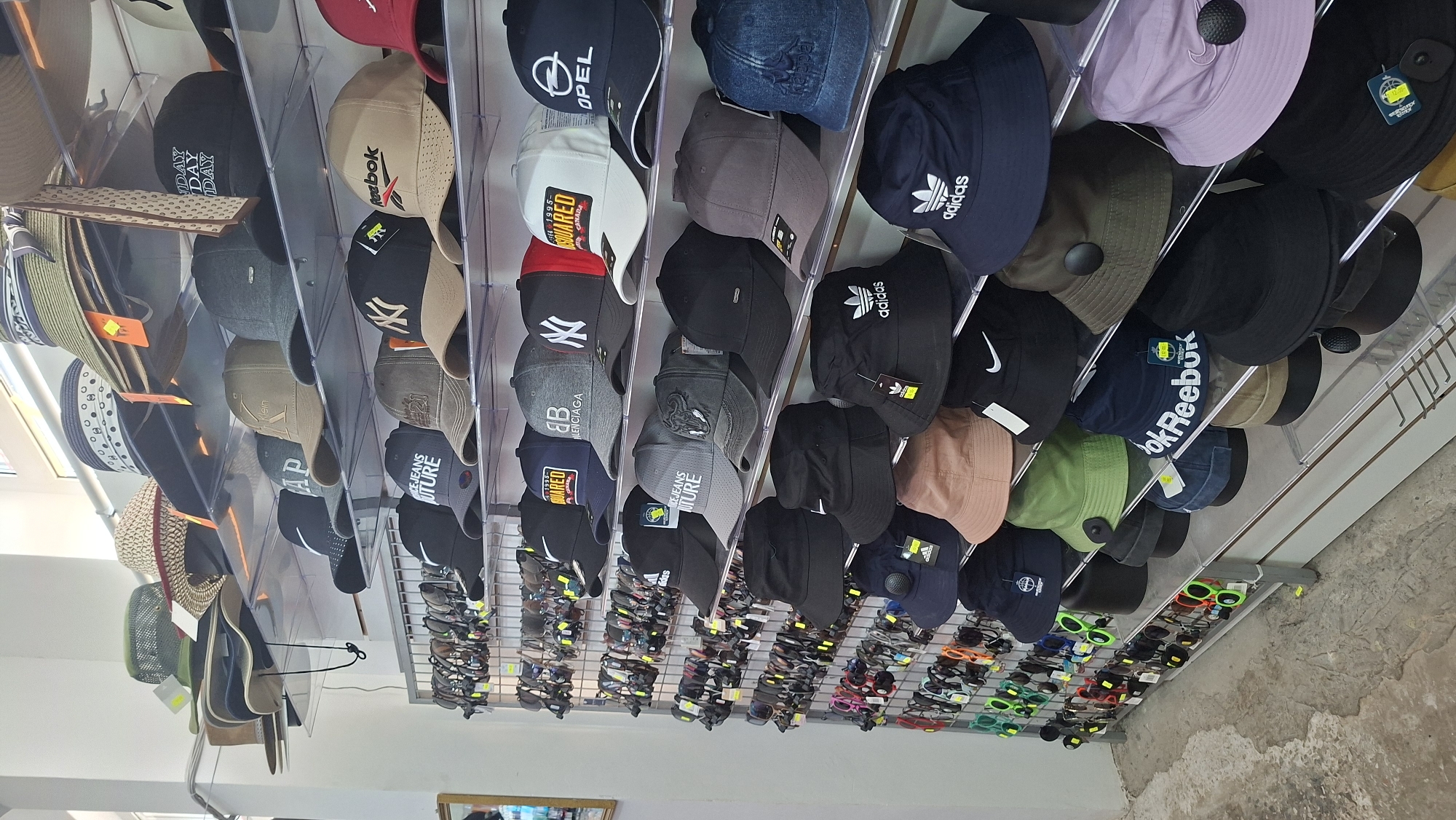 A retail display of hats and sunglasses is shown in a store. The hats are arranged on shelves, with different styles and brands visible, including baseball caps and bucket hats. Sunglasses are displayed on racks to the left of the hats. The environment appears to be a well-lit indoor space with white walls and shelving units. There are no people visible in the image. The city of Myadzyel, Belarus is not evident in the provided image.