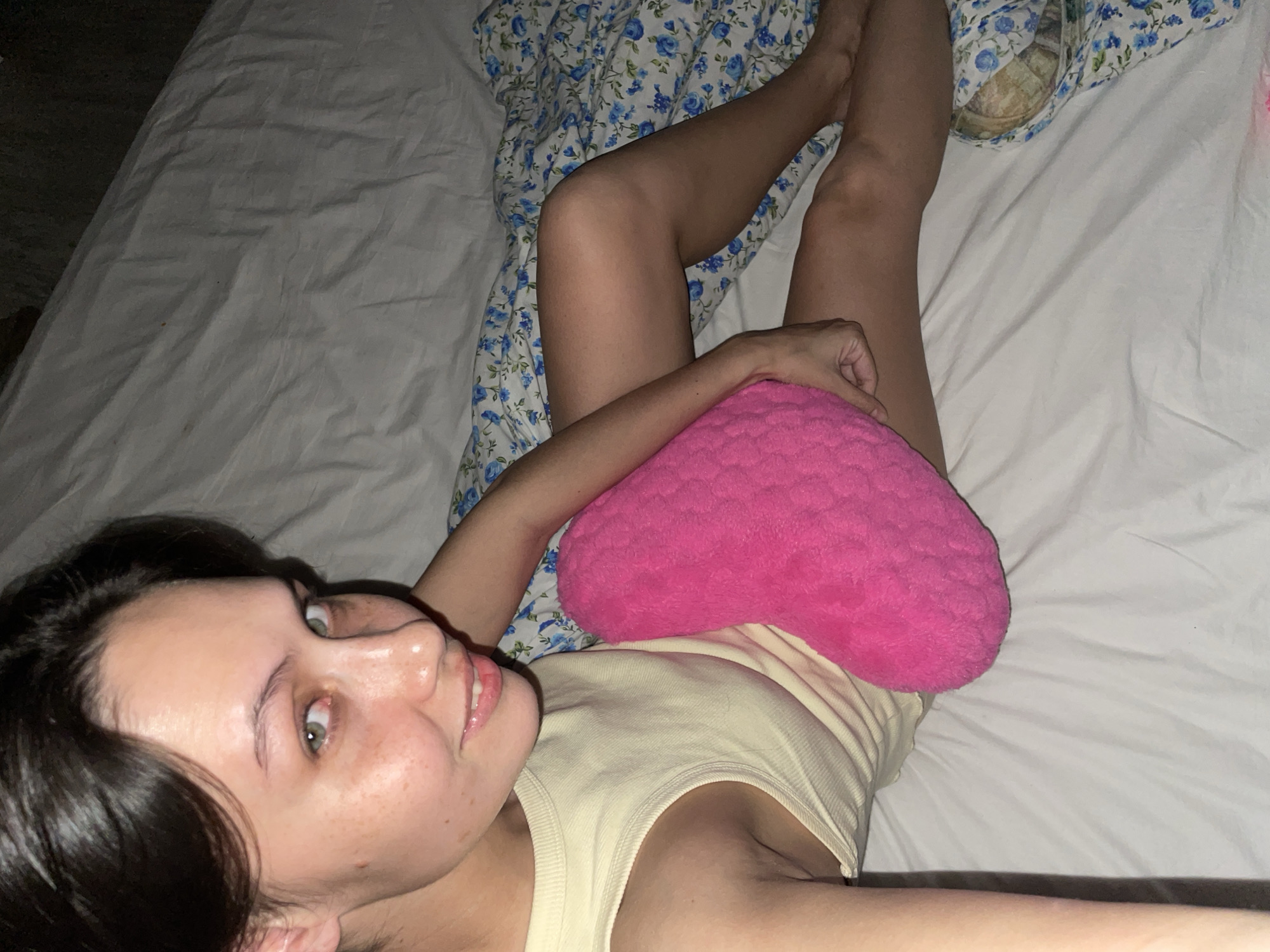 A woman with dark hair and light skin is lying on a bed with white sheets. She is wearing a light yellow tank top and is holding a bright pink heart-shaped pillow. Her legs are visible, with a patterned blanket behind them. No information about Izhevsk, Russia, is discernible from the image.