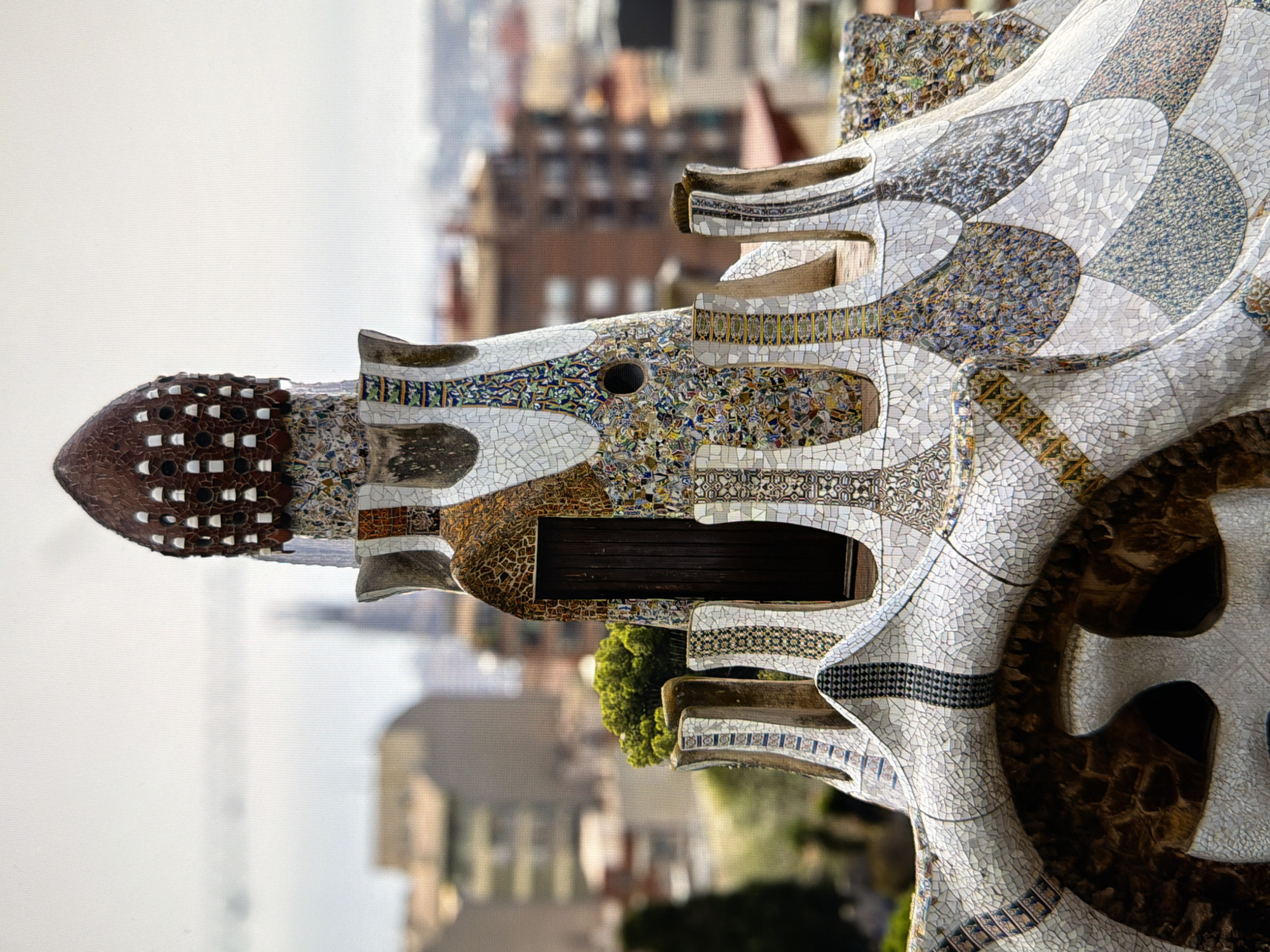 The image displays a close-up of a mosaic-covered turret, possibly part of a larger architectural structure. The turret features intricate patterns and vibrant colors created by broken ceramic tiles. A dark wooden door is visible on the side of the turret. In the background, a blurred cityscape with buildings and trees suggests an urban environment. The sky is overcast and appears pale. The architectural style, characterized by its whimsical and organic forms, is reminiscent of Antoni Gaudí's work, a prominent architect known for his unique style in Barcelona, Spain. This suggests the location is likely Barcelona, not Kyiv, Ukraine. There are no people visible in the image.