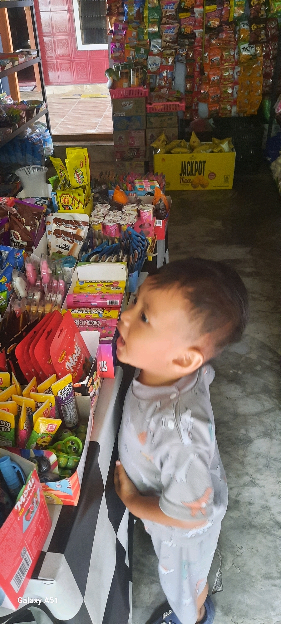 In Ploso, Indonesia, a young child stands in front of a market stall filled with snacks. The child, wearing a grey onesie with dinosaur prints, looks to the right with an open mouth, as if in awe or anticipation. The stall displays a variety of brightly packaged candies and snacks, including red packets labeled "Modi 3D Fruit Salad," tubes of "Choco Tube," and boxes of "Chocolate Beans." Behind the child, shelves are packed with more colorful snack bags, and a large yellow box with the word "JACKPOT" is visible. The market floor appears to be concrete. The overall scene depicts a child captivated by the abundance of treats in a local Indonesian market.