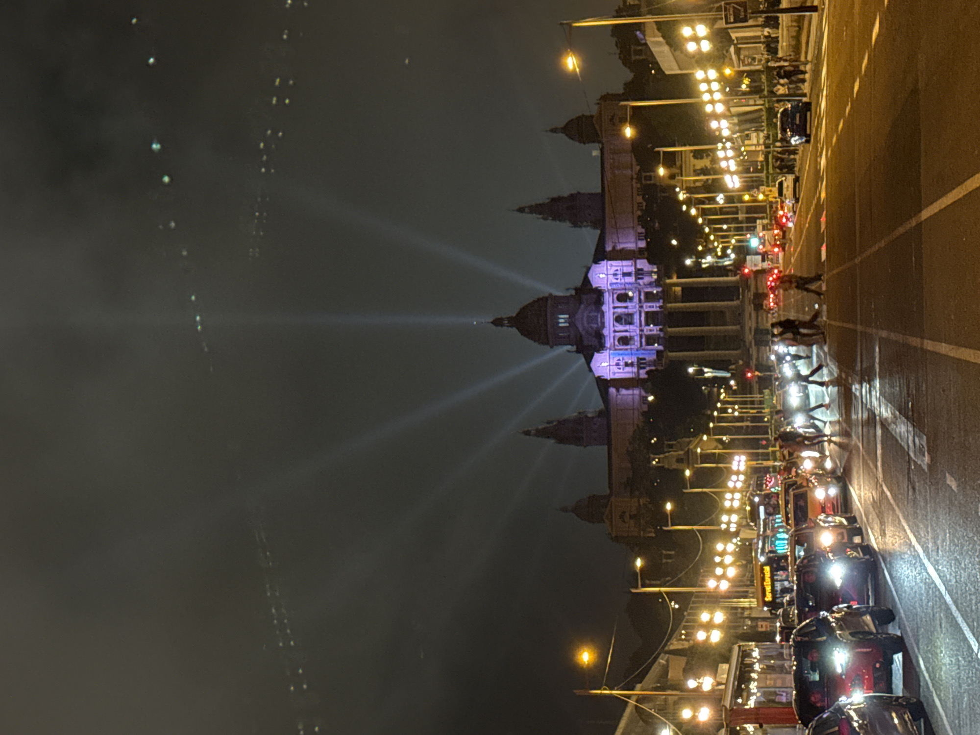 The image depicts the Palau Nacional in Barcelona, Spain, illuminated with light beams in the night sky. The Palau Nacional is situated at the end of Avinguda de la Reina Maria Cristina. This avenue is shown filled with traffic. Pedestrians are walking on the road. Streetlights line the avenue, creating a brightly lit pathway towards the palace. The sky is dark, with streaks of light, and the overall scene is a vibrant display of urban activity at night.
