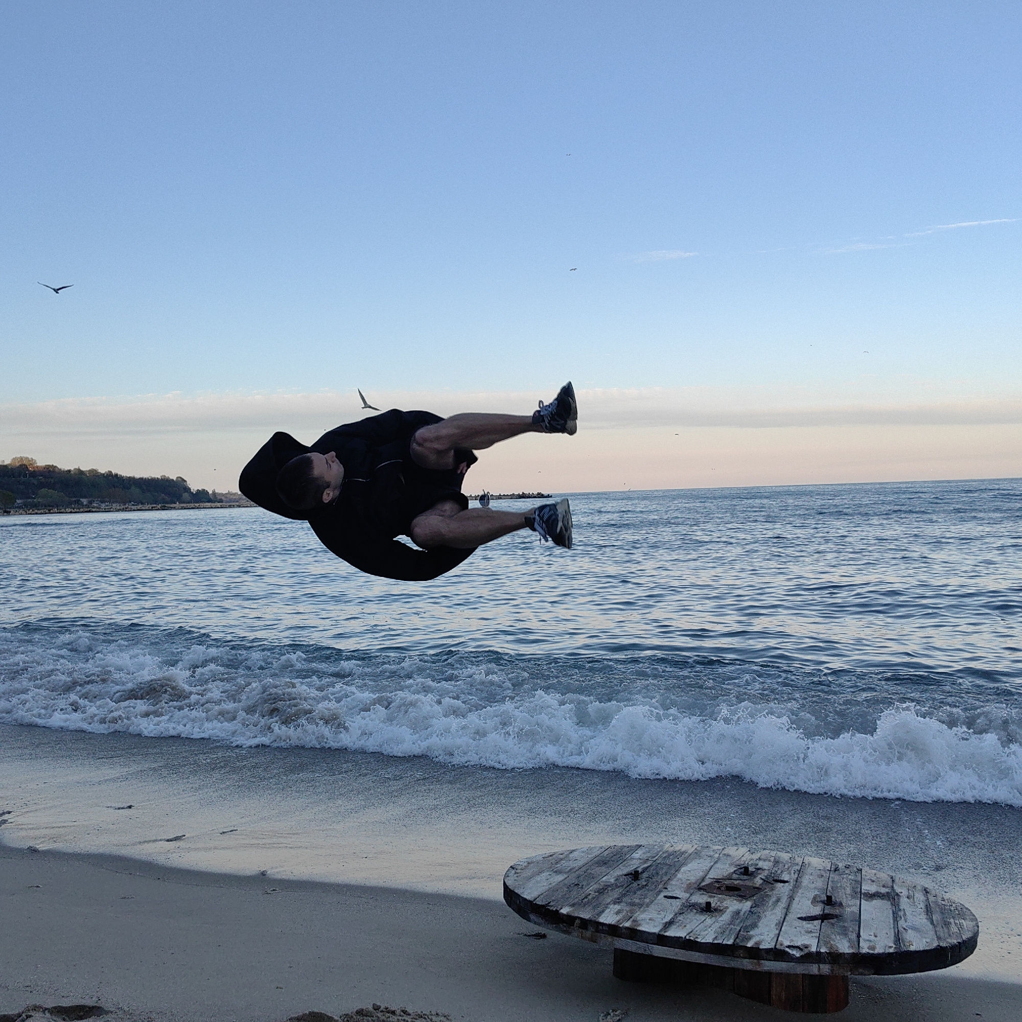 The image captures a man performing a mid-air acrobatic flip near the Black Sea coast in Varna, Bulgaria. The background shows a coastal landscape with a beach, waves, and a horizon line where the sea meets the sky. The sky transitions from blue to a soft gradient of light pink and white. Several birds are visible in the sky. On the beach, there's a weathered, round, wooden cable spool in the lower right corner. The man is wearing dark clothing and athletic shoes, positioned horizontally in the air, seemingly in the middle of a backflip or similar acrobatic movement. The surrounding beach has foamy waves crashing onto the shore. In the distance, a strip of land with some vegetation is visible.