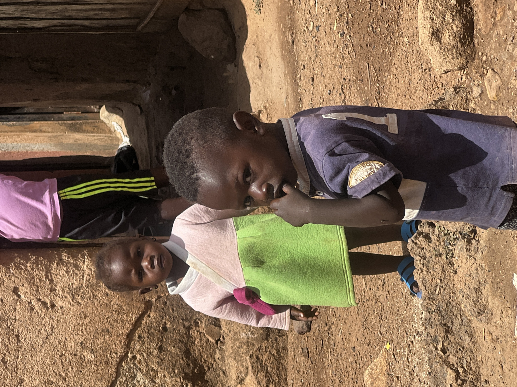 Two young children stand on a dirt surface in Gura Top, Nigeria. One child wears a pink and green dress with a matching bag. The other wears a blue shirt with the number "1" on the sleeve and blue sandals. Another person wearing black pants with yellow stripes and a pink shirt is partially visible in the background. The setting appears to be an impoverished area. The light suggests it is daytime.