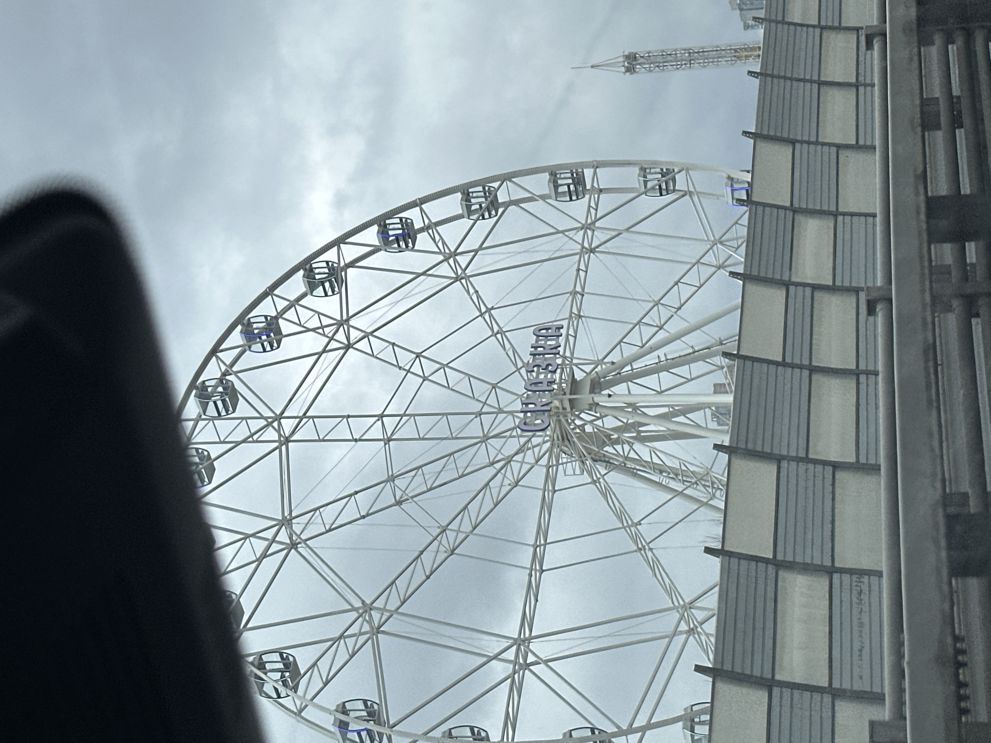 The image depicts a partial view of the "Sun of Moscow" Ferris wheel, located in Moscow, Russia. The wheel's white structure with enclosed passenger cars is visible against an overcast sky. Adjacent to the wheel is a building with a gray and white facade and metal support structures. In the background, there is a glimpse of a construction crane. The foreground includes part of a dark object, possibly a vehicle interior, obscuring the lower left portion of the frame.