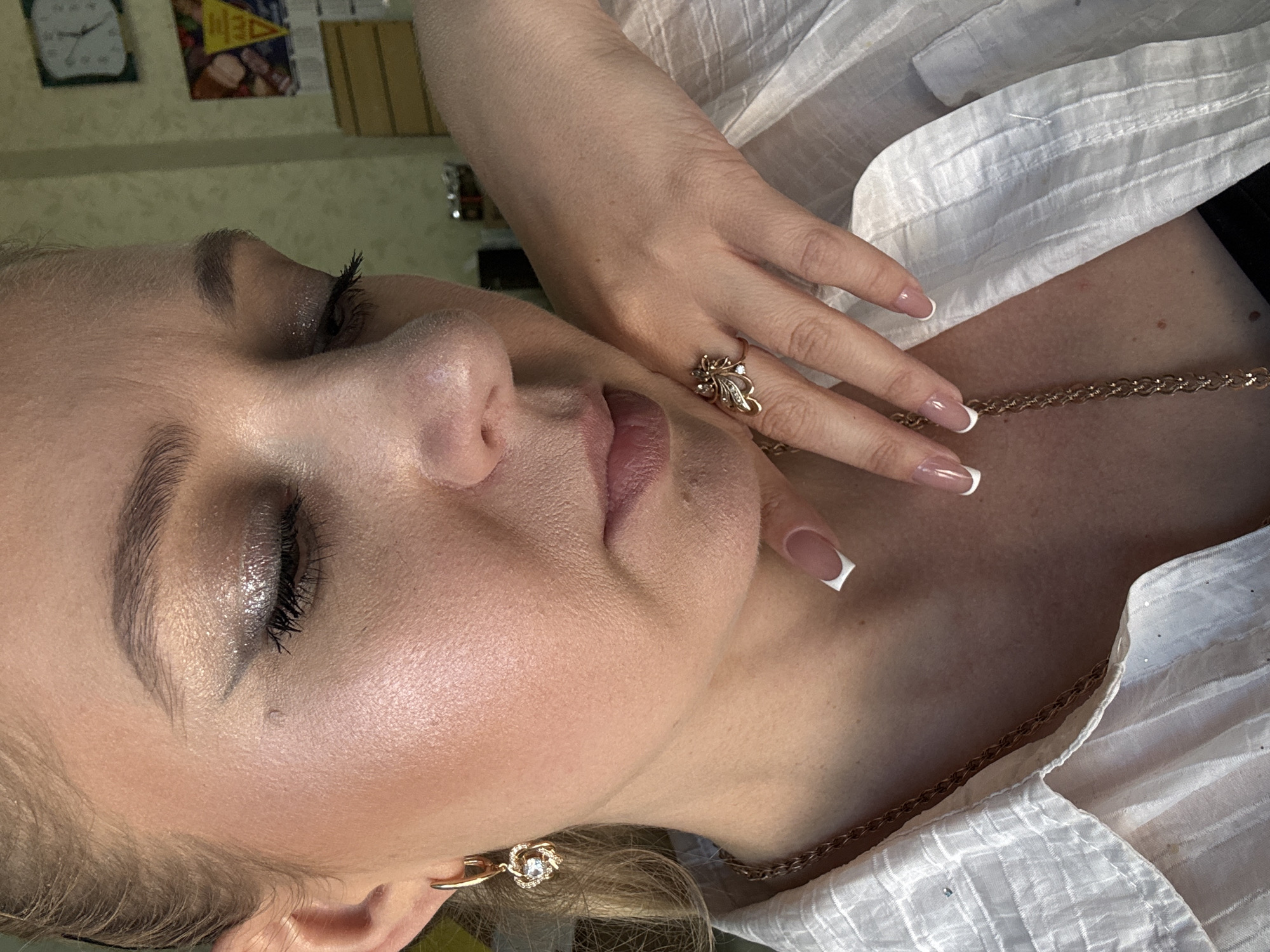 A close-up, medium shot shows a fair-skinned woman from the chest up, angled in profile with her eyes closed and head tilted down. She is wearing a white, textured blouse and several pieces of gold jewelry: a dangling heart-shaped earring with a clear stone, a ring with a floral design and small stones on her right hand, and a thick gold chain necklace. Her nails are manicured with a French tip design. Her makeup includes dark eyeliner, mascara, and shimmery eyeshadow. The background is blurred, with hints of a wall with a clock and some framed pictures, suggesting an indoor setting.