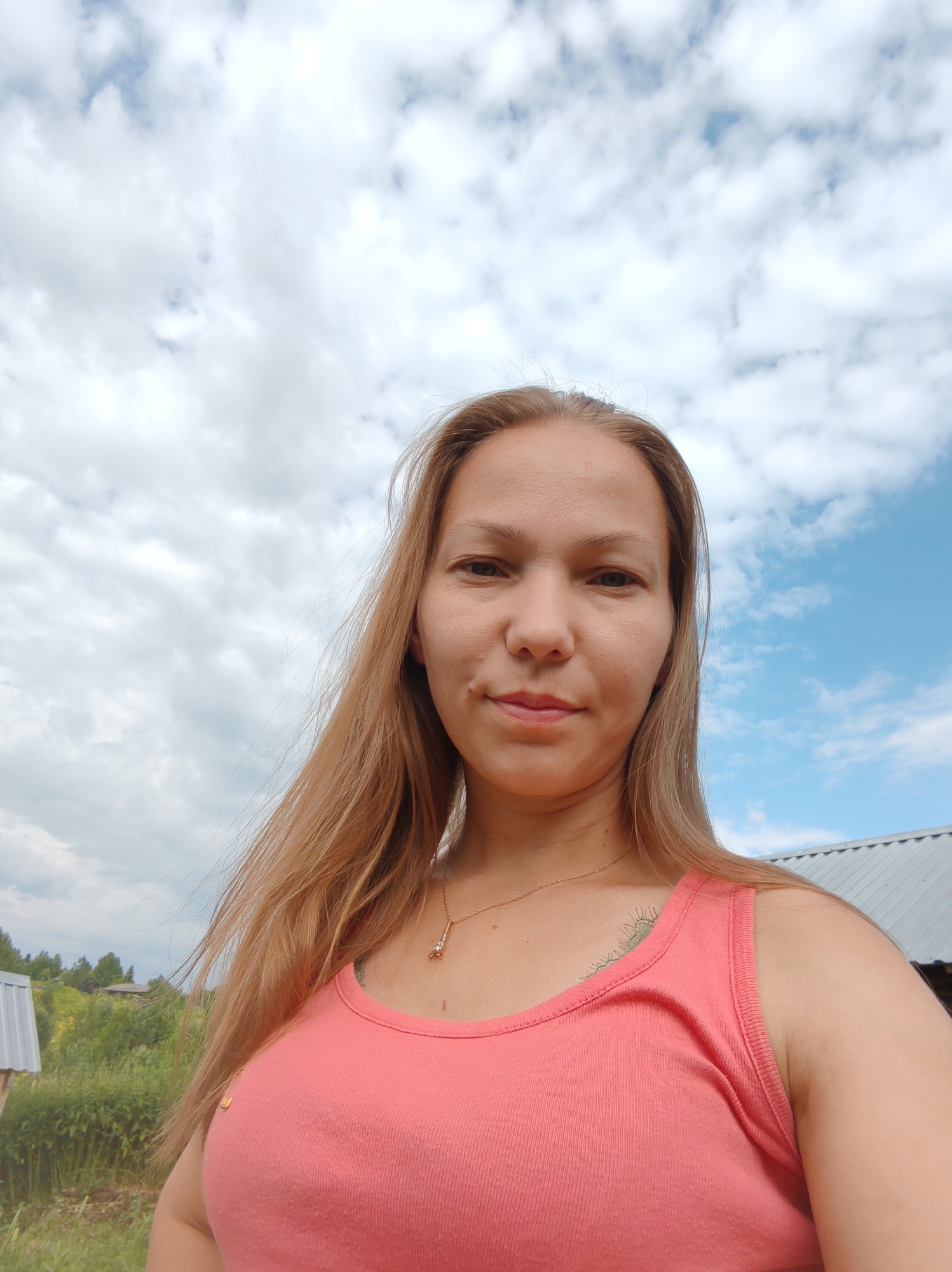 A medium shot from below shows a woman with blonde hair and fair skin, wearing a coral-colored tank top, standing outdoors under a partly cloudy sky. Her hair is pulled back slightly to reveal her face, which has a neutral expression. She is wearing a delicate gold necklace with a pendant. Behind her, a rural landscape is visible, with green foliage, trees, and a rustic building with a metal roof. The sky is a mix of blue and white clouds. The location is the outskirts of Chukhlem, Russia.
