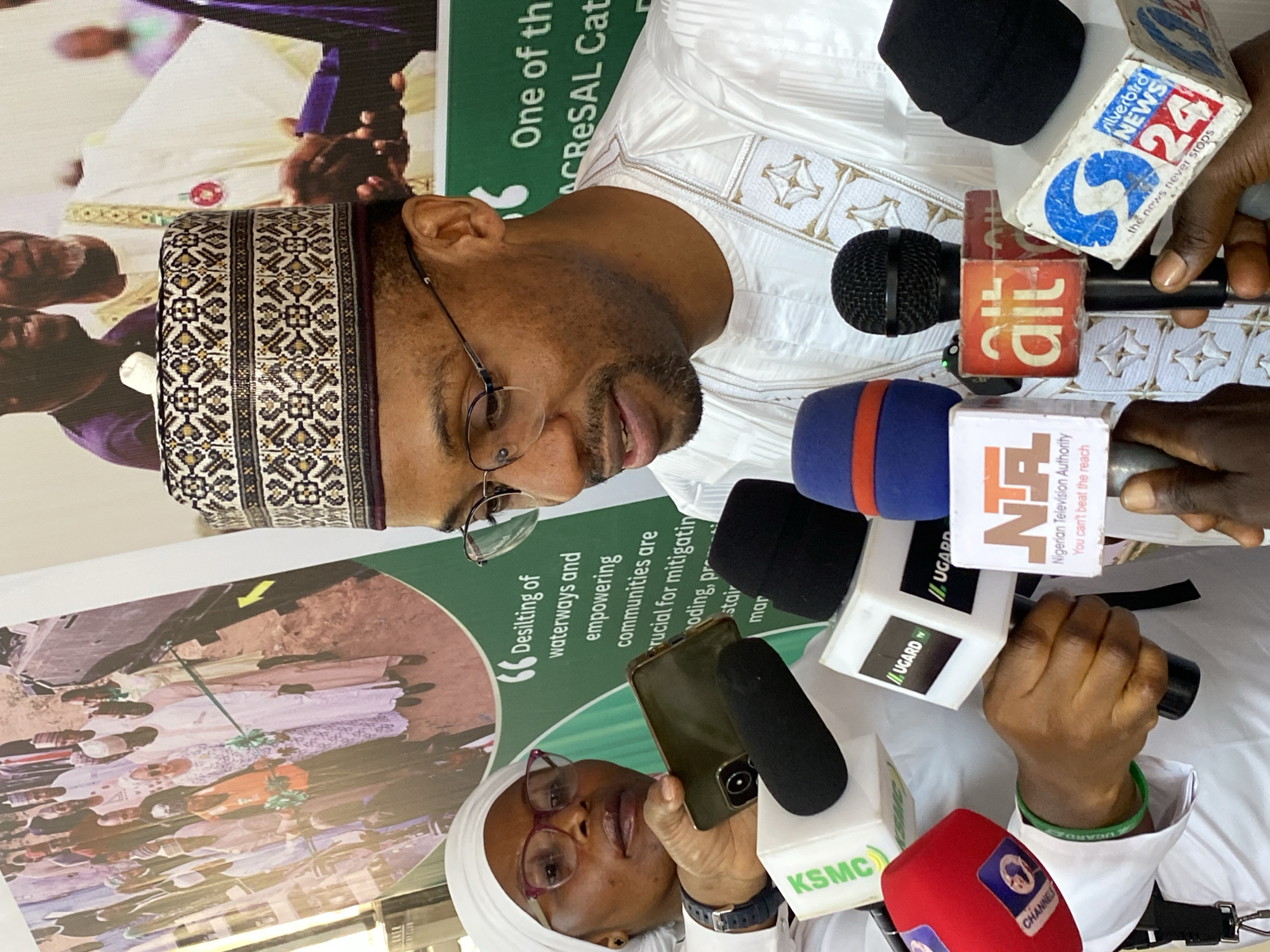 This image captures a media briefing or press conference held in Kwarau, Nigeria. The central figure is a man, likely a dignitary or official, wearing traditional Nigerian attire consisting of a white embroidered agbada and a patterned cap. He also wears glasses and has a neatly trimmed beard. He is seen speaking into a cluster of microphones, indicating an official statement or address to the press.
Surrounding the speaker are numerous microphones held by various individuals, representing different media organizations. Visible logos on the microphone covers include "silverbird NEWS 24" with the slogan "the news never stops," "ait," "NTA Nigerian Television Authority" with the slogan "You can't beat the reach," "UGARD TV," "KSMC," and "CHANNELS." In the foreground, a woman wearing a white hijab and glasses is partially visible, holding a smartphone, seemingly recording the event. One hand of a person holding a microphone wears a green wristband with "UGARDW" visible.
Behind the speaker, a large banner is displayed, featuring text that reads: "Desilting of waterways and empowering communities are crucial for mitigatin oding, pr stair mar" and "One of th ACRESAL Cat." This suggests the event is related to environmental or community development projects. Another section of the banner shows a large circular image of several people, possibly indicating community engagement or project beneficiaries. The lighting suggests it is daytime. The overall scene conveys a formal and informative gathering, characteristic of an official announcement or public address.