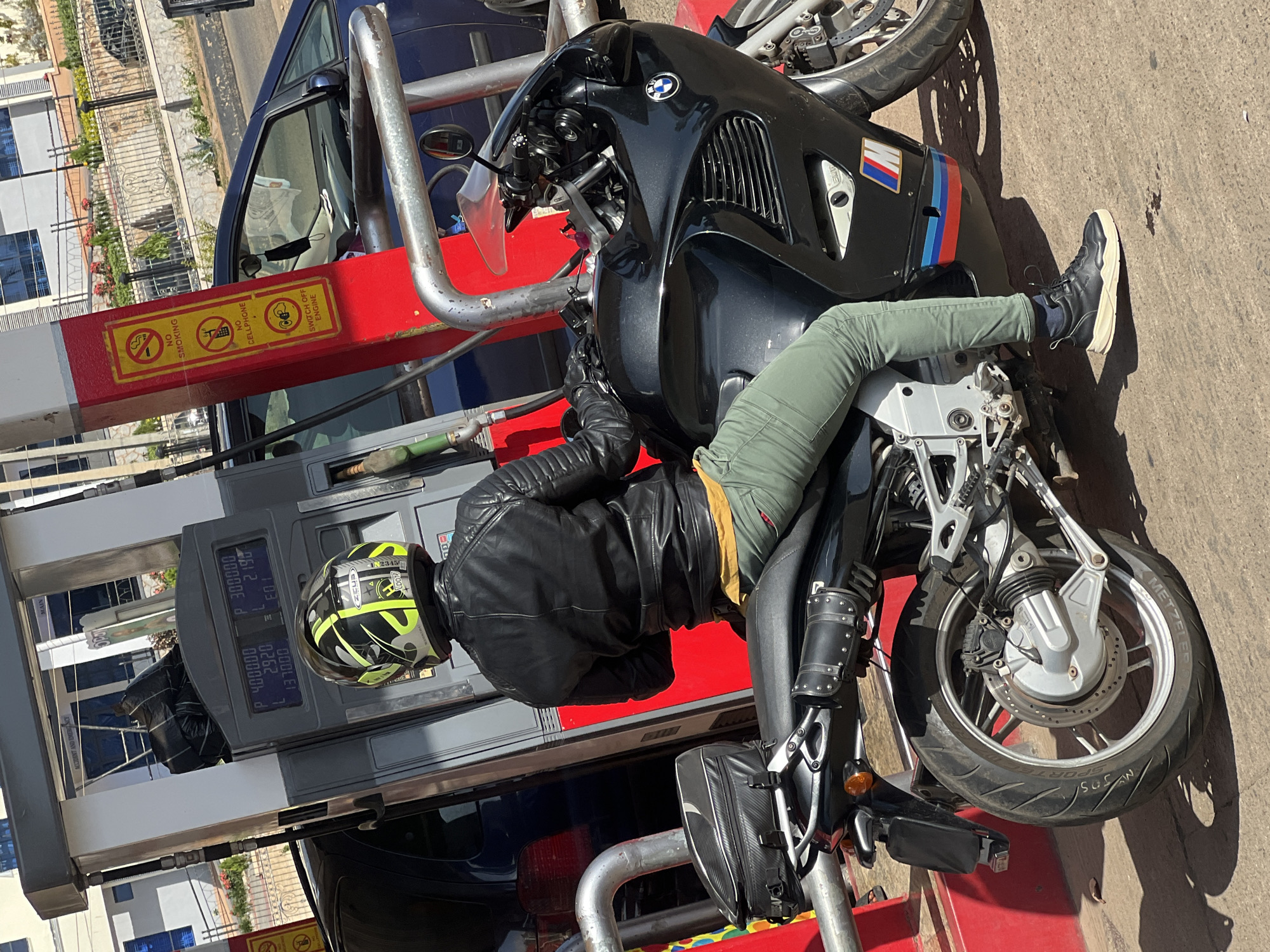 A motorcyclist in Gura Top, Nigeria, is refueling at a gas station. The person is wearing a black leather jacket, light green pants, black shoes, and a yellow/black helmet. They are seated on a black BMW motorcycle next to a gas pump that is currently dispensing fuel, indicated by the digital display showing "P 4000.00," "L 2.920," and "1370.00." The gas pump has red panels with yellow warning signs including "No Smoking," "No Cellphone," and "Switch Off Engine." A blue car is parked adjacent to the gas pump.
