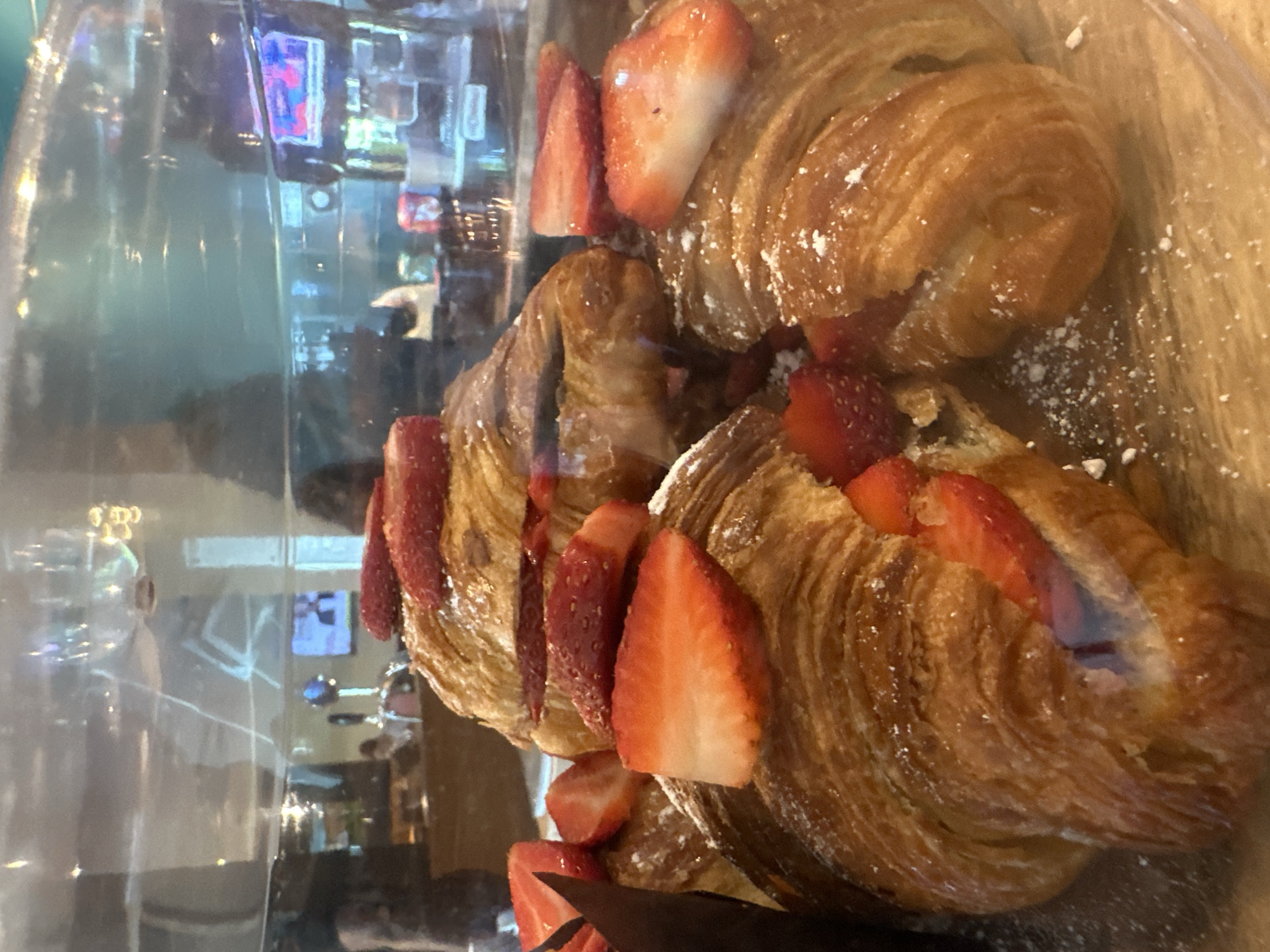 Two croissants filled with sliced strawberries and dusted with powdered sugar are displayed in a glass case. Reflections in the glass obscure the background, which appears to be a building with televisions and people. The location is likely London, United Kingdom, but this cannot be confirmed with certainty from the image.