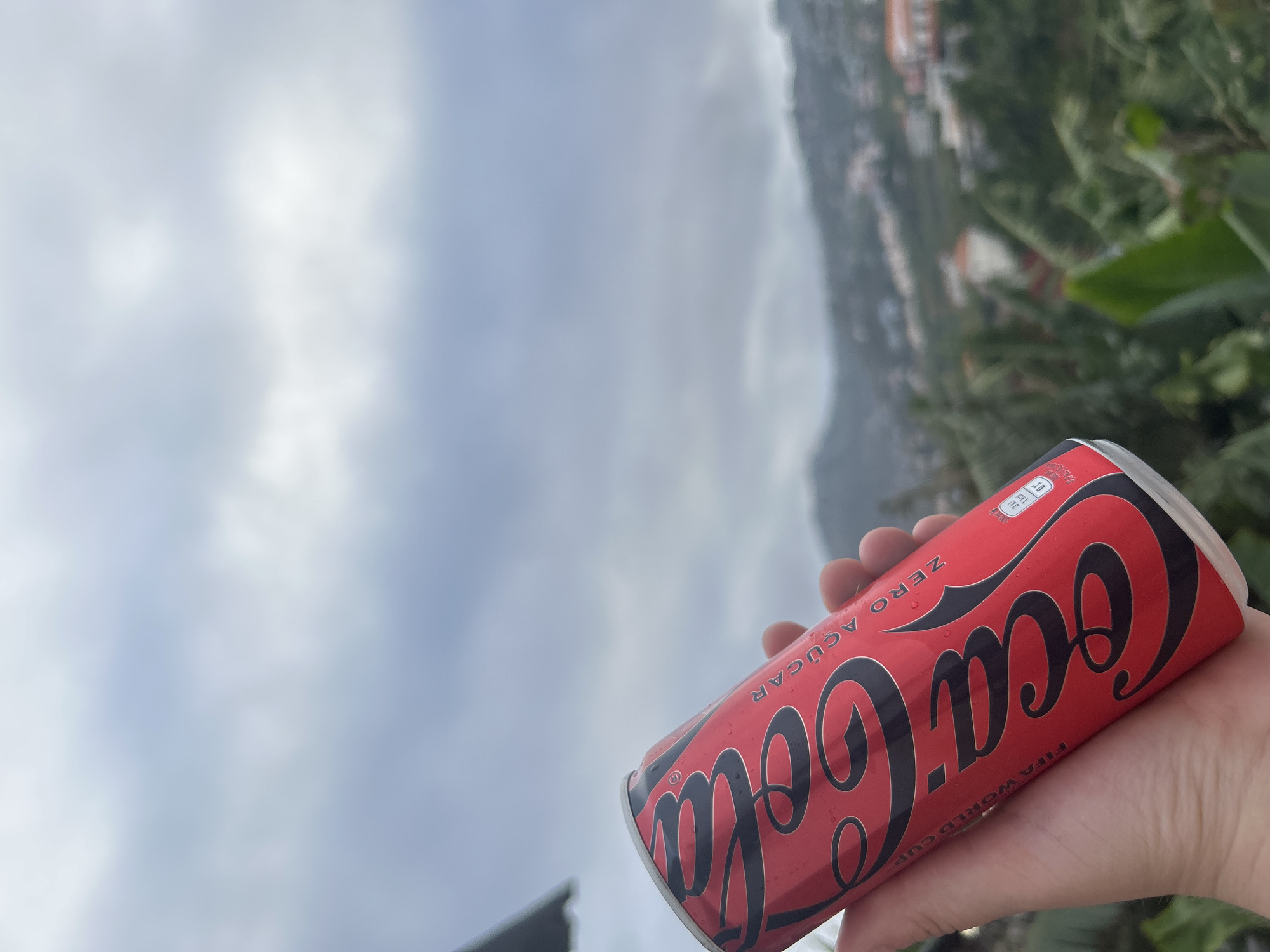 A person holds a can of Coca-Cola Zero Sugar in their right hand. The can is red with the Coca-Cola logo in black. "ZERO AÇUCAR" is printed on the can. Water droplets are visible on the can's surface. The background features a cloudy sky and part of the city of Câmara de Lobos, Portugal, with buildings and green foliage visible on the hillside. The camera angle looks upward toward the sky and city.