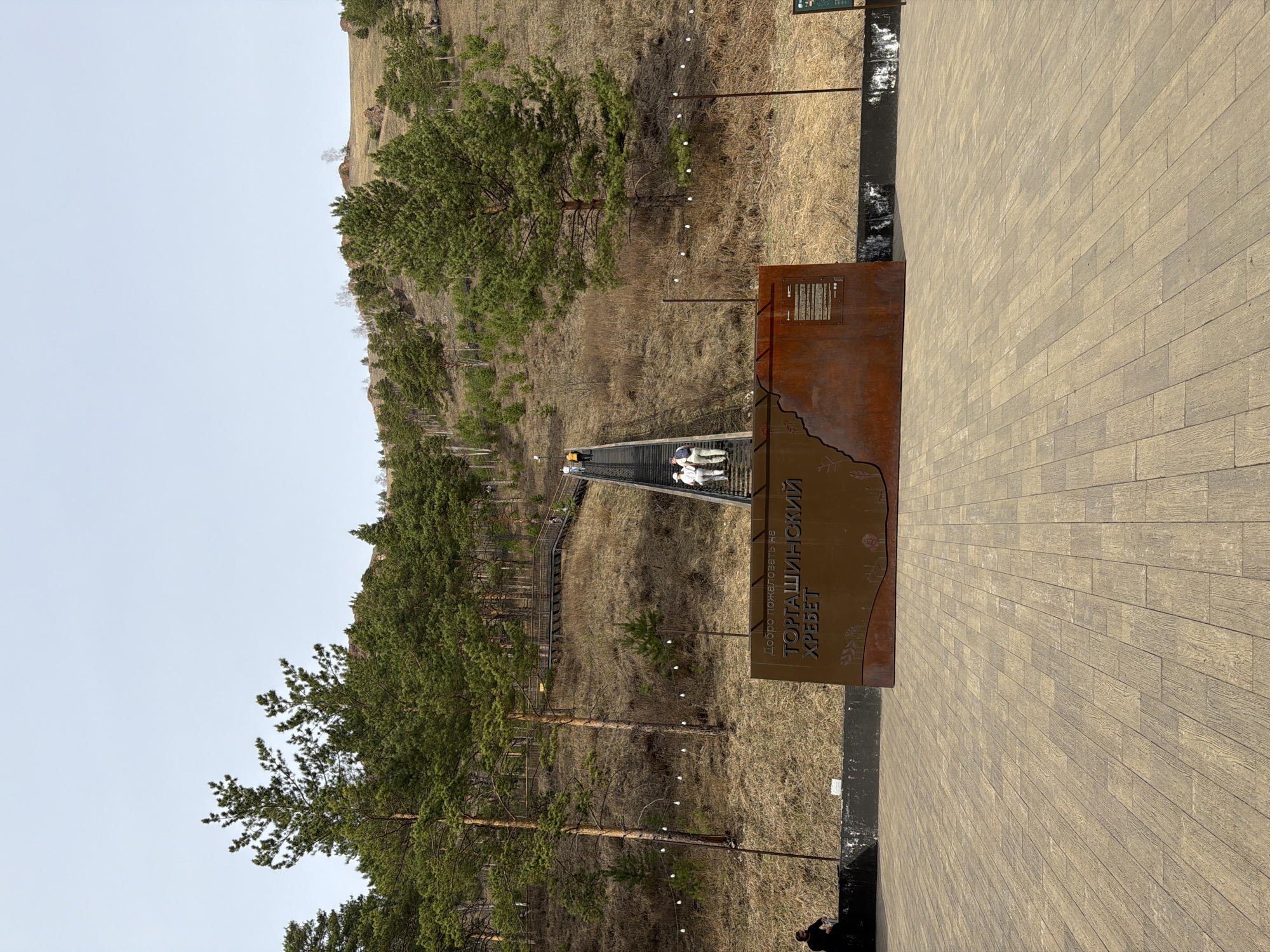 The image presents an outdoor scene at the Torgashinsky Ridge in Krasnoyarsk, Russia. In the foreground, a wide, light-colored paved area, resembling wood planks, provides a solid ground surface. To the left on this platform stands a prominent, large, rusted metal sign. This sign features a cutout silhouette at its base and bears clear Russian text.
From this paved area, a long, multi-tiered wooden or metal staircase and boardwalk system extends upwards, winding along a hillside into the distance. Several people are visible on this ascending path, with two individuals in light-colored clothing clearly making their way up the stairs. The hillside is sparsely vegetated with dry, light brown grass and numerous green pine trees scattered across its slopes. Along the path's edge, a series of poles equipped with small white globe lights are installed.
The sky in the upper portion of the frame is clear and light, indicating daytime conditions. The dry vegetation suggests a season like late spring or early autumn. The visible text on the large rusty sign reads: "ДОБРО ПОЖАЛОВАТЬ НА ТОРГАШИНСКИЙ ХРЕБЕТ," translating to "Welcome to Torgashinsky Ridge." The scene captures people engaging in a leisure activity, likely hiking or sightseeing, within a natural, developed recreational area.
