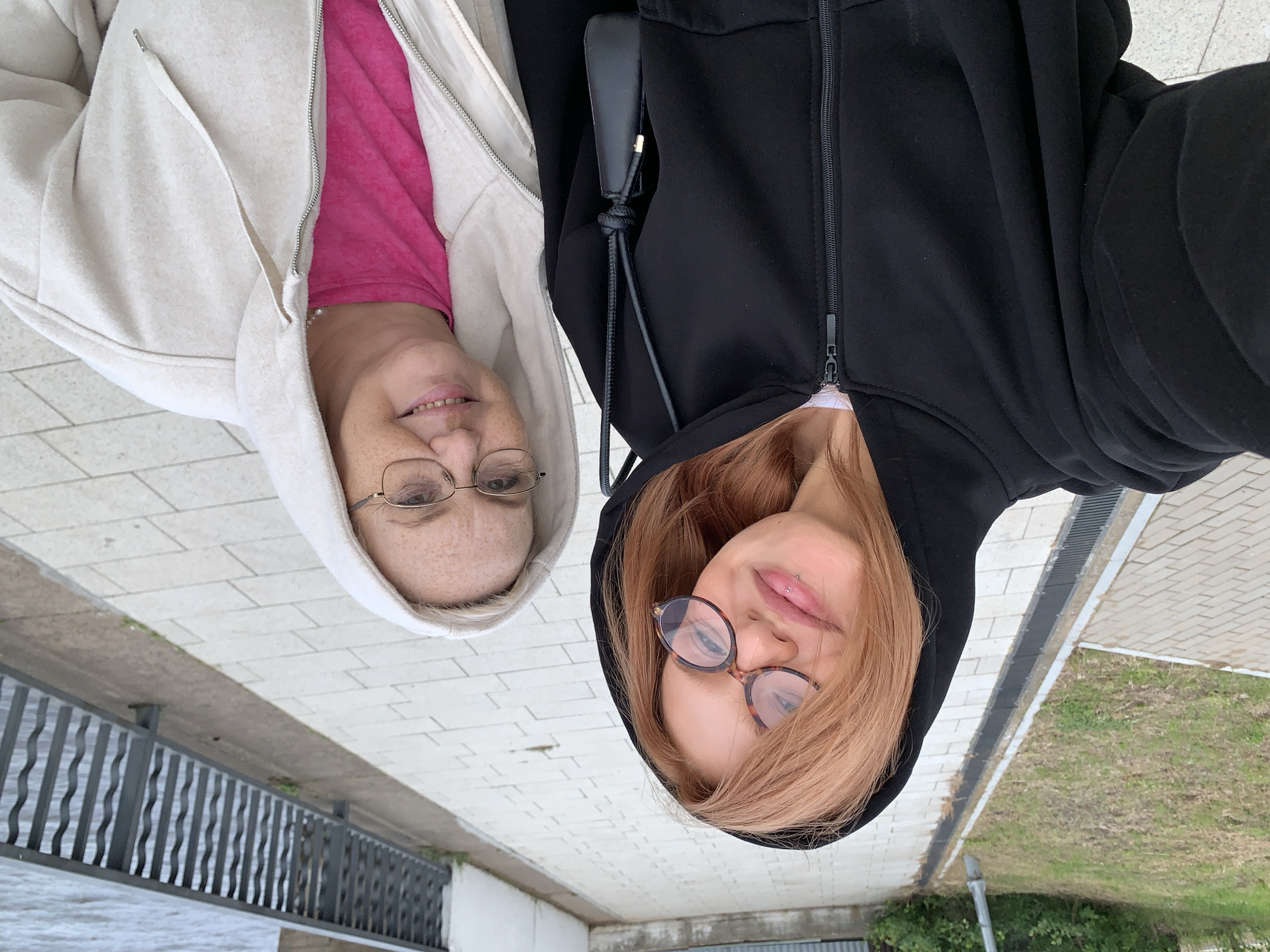 A selfie-style photograph depicts two smiling women posing outdoors on a paved walkway in Chaikovsky, Russia. The woman in the foreground on the left has light reddish-brown hair, wears tortoise-shell glasses, and a black zip-up hooded jacket with a white layer underneath. A labret piercing is visible below her lower lip, and she is holding a small black shoulder bag with a knotted strap. The woman on the right, partially visible, has fair skin with noticeable freckles, wears gold-rimmed glasses, and an off-white or light beige zip-up hooded jacket over a bright pink t-shirt, with a pearl necklace around her neck. Both women are smiling directly at the camera.
The environment includes a light-colored paved pathway composed of rectangular blocks. A linear drain with a metal grate separates the pathway from a strip of grass and further green vegetation, including bushes and trees, on the left. To the right, a dark grey metal railing with decorative wavy vertical bars extends along the edge of the walkway, overlooking a body of water with visible ripples, indicative of a river or large lake. An overcast sky provides diffused lighting for the scene.