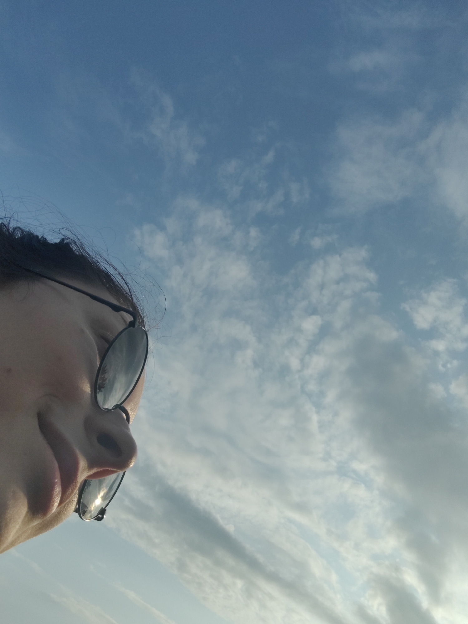 A close-up, low-angle shot captures the side profile of a person wearing dark-rimmed sunglasses. Their dark hair is visible at the top of the frame. The person's face is tilted upwards, looking towards a bright, cloudy blue sky. The clouds are wispy and scattered, suggesting an outdoor setting, possibly during daytime. The reflection of trees and the sky is visible in the lenses of the sunglasses. The overall scene conveys a relaxed, outdoor atmosphere. The image was taken in Ермолаево, Russia.