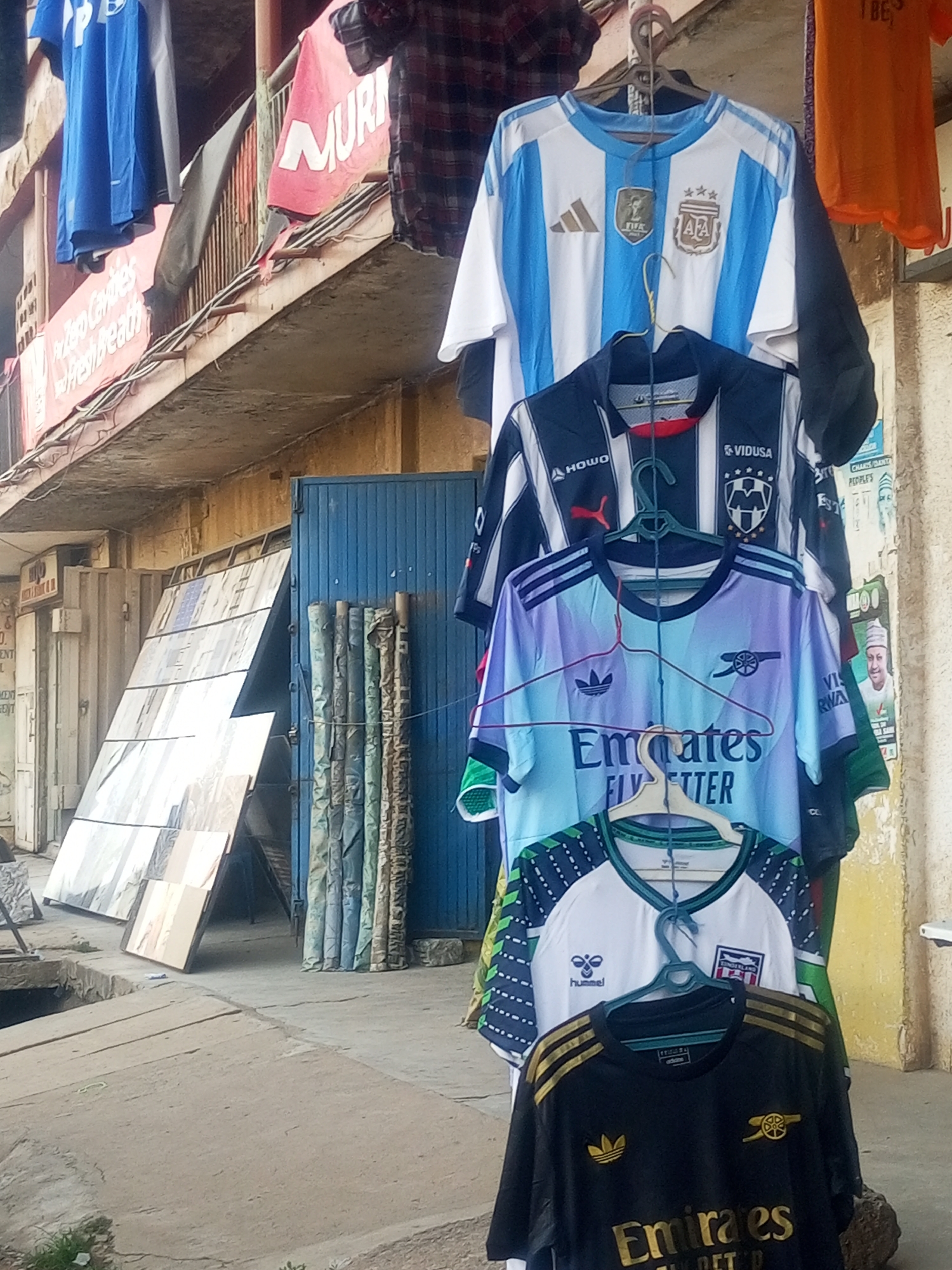 The image shows a street scene in Maiburiji, Nigeria, with soccer jerseys hanging for sale outside a shop. The jerseys represent teams like Argentina and Arsenal. The shop front has a corrugated metal door and various signs and posters are visible on the walls. There are also metal sheets leaning against the shop and rolls of material stacked to the side. The street appears to be a commercial area with other shops and buildings visible in the background.