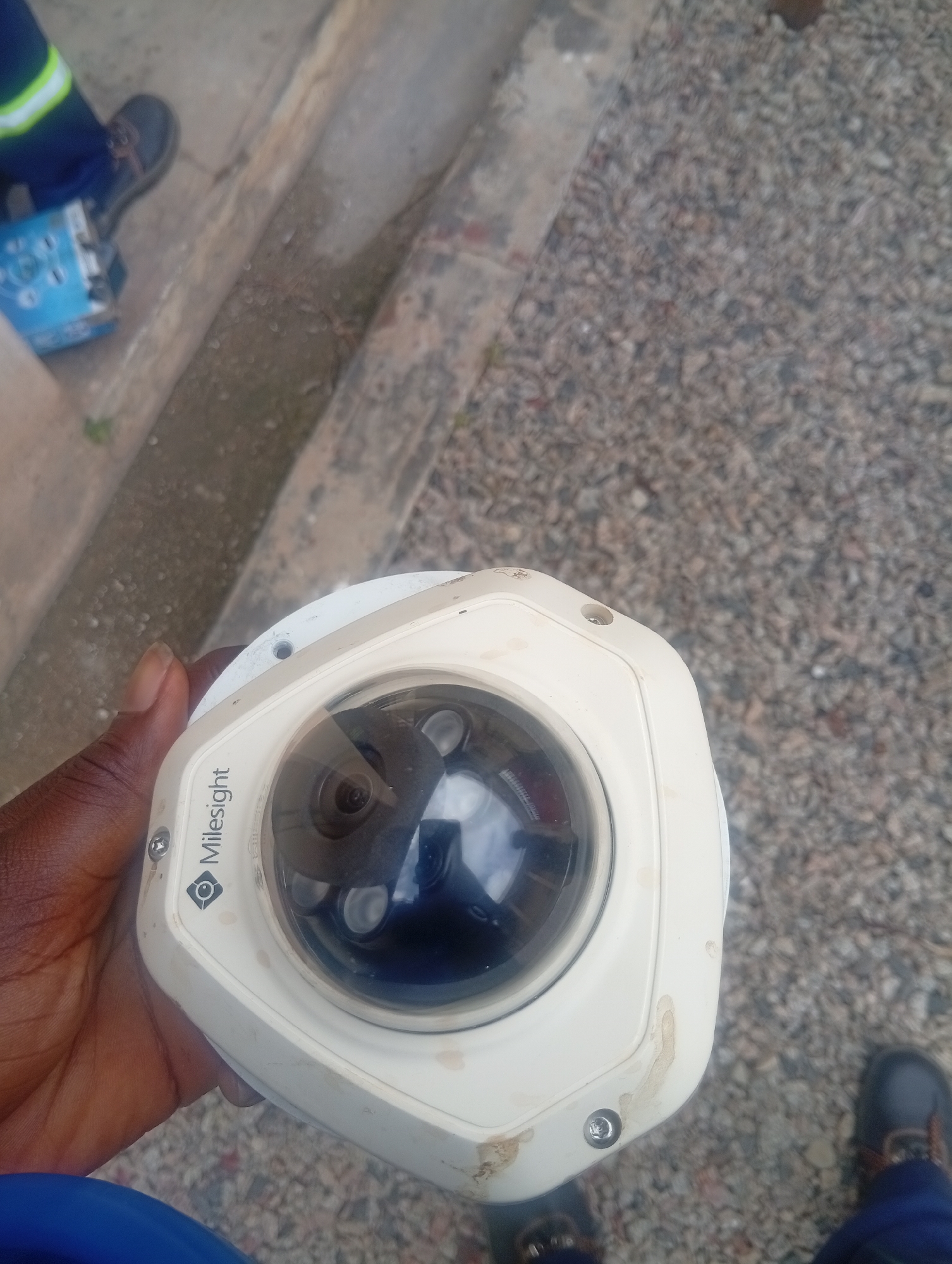 In Shiroro, Nigeria, a close-up shot shows a person's hand holding a white, dome-shaped security camera with the brand name "Milesight" clearly visible. The camera appears to be made of plastic and has a reflective lens, hinting at its surveillance capabilities. The background is a mix of rough, grey gravel and a concrete walkway, suggesting an outdoor urban or semi-urban setting. A portion of a blue and yellow safety garment and a dark-colored shoe are visible in the upper left of the frame, implying the presence of a worker. The scene is well-lit, likely by natural daylight.
