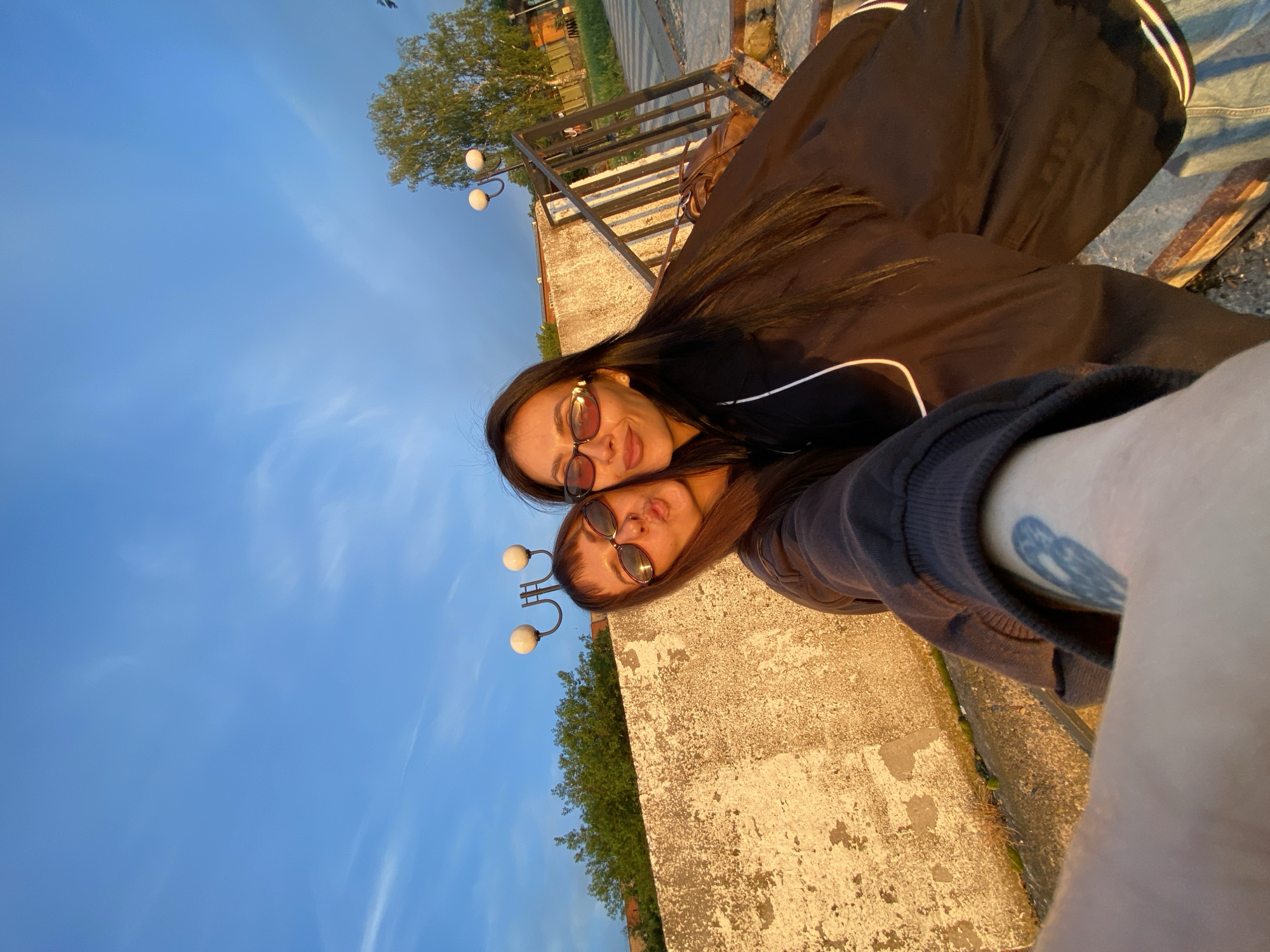 Two women pose for a selfie outdoors. They are positioned close together, wearing sunglasses and jackets. A concrete barrier is visible behind them, with vegetation and lampposts in the background. The sky is blue with light clouds. A metal railing and steps are present in the background. The scene is illuminated by warm, golden sunlight, suggesting it's either sunrise or sunset. The presence of lampposts and building architecture may indicate it is near the embankment of the Votkinsk Pond in the city of Votkinsk, Russia.